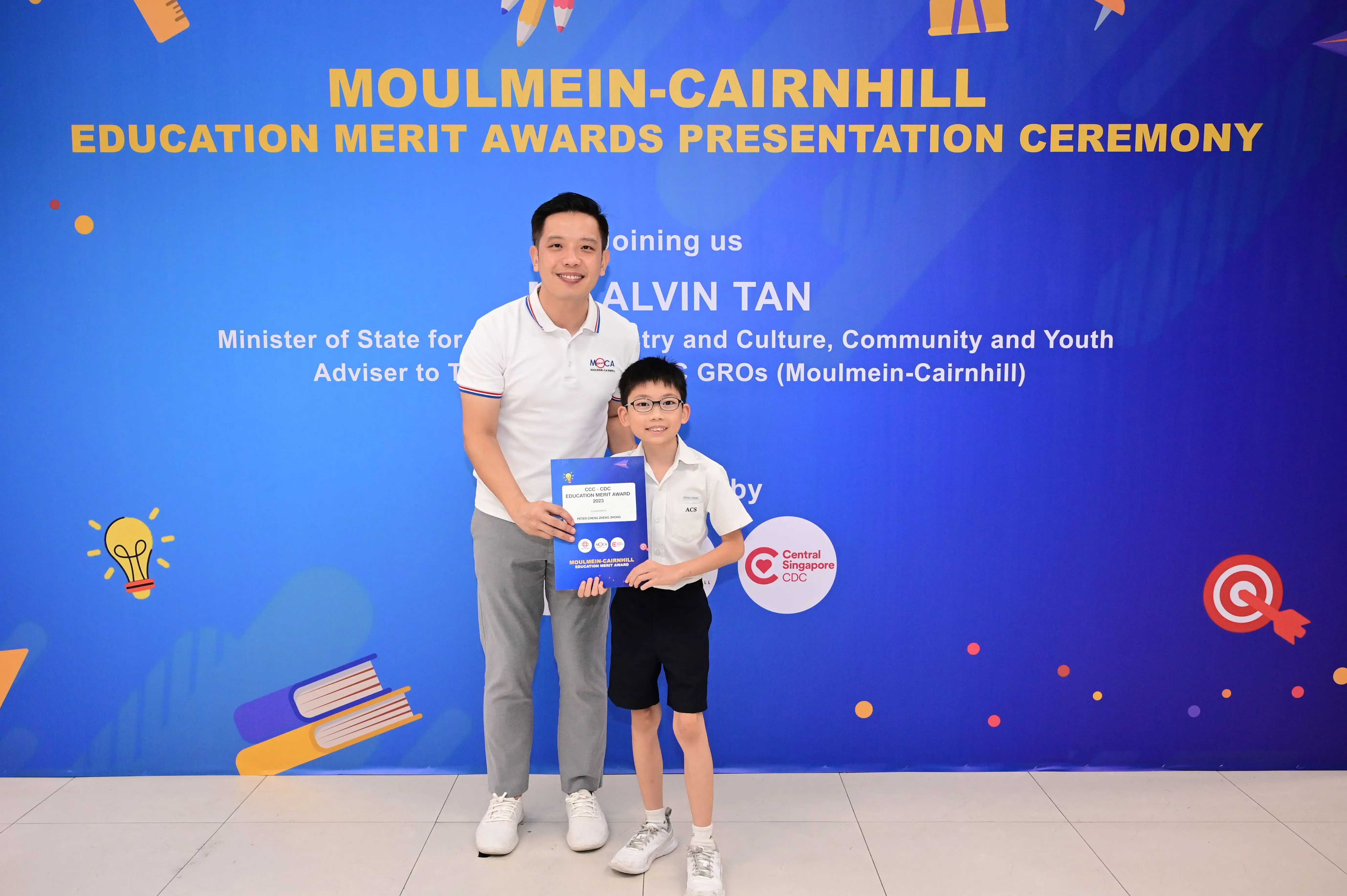 A man and a young boy holding a blue award certificate stand before a Moulmein-Cairnhill Education Merit Awards banner.