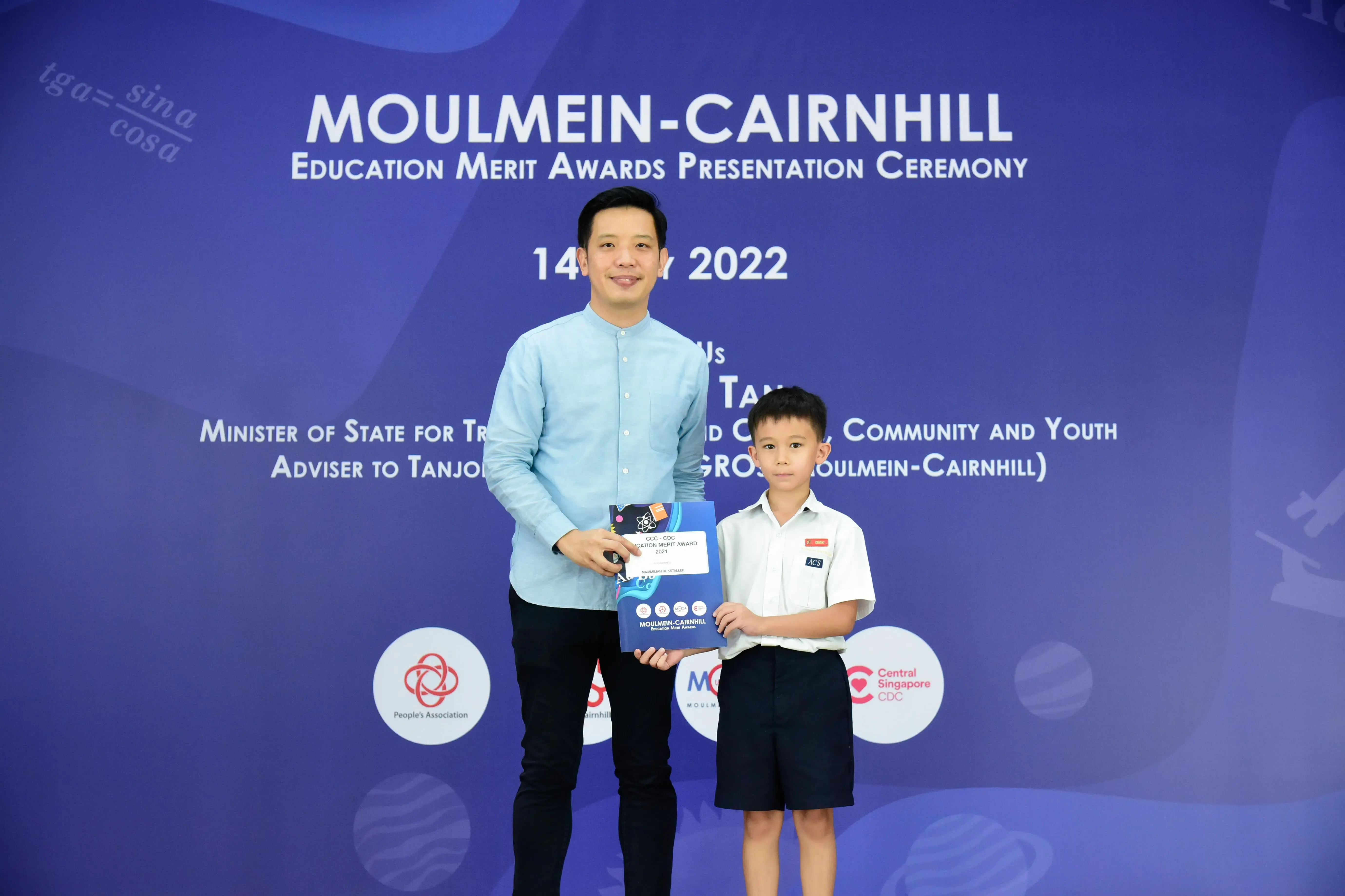 A man and a young boy in school uniform hold an award in front of a Moulmein-Cairnhill Education Merit Awards banner.
