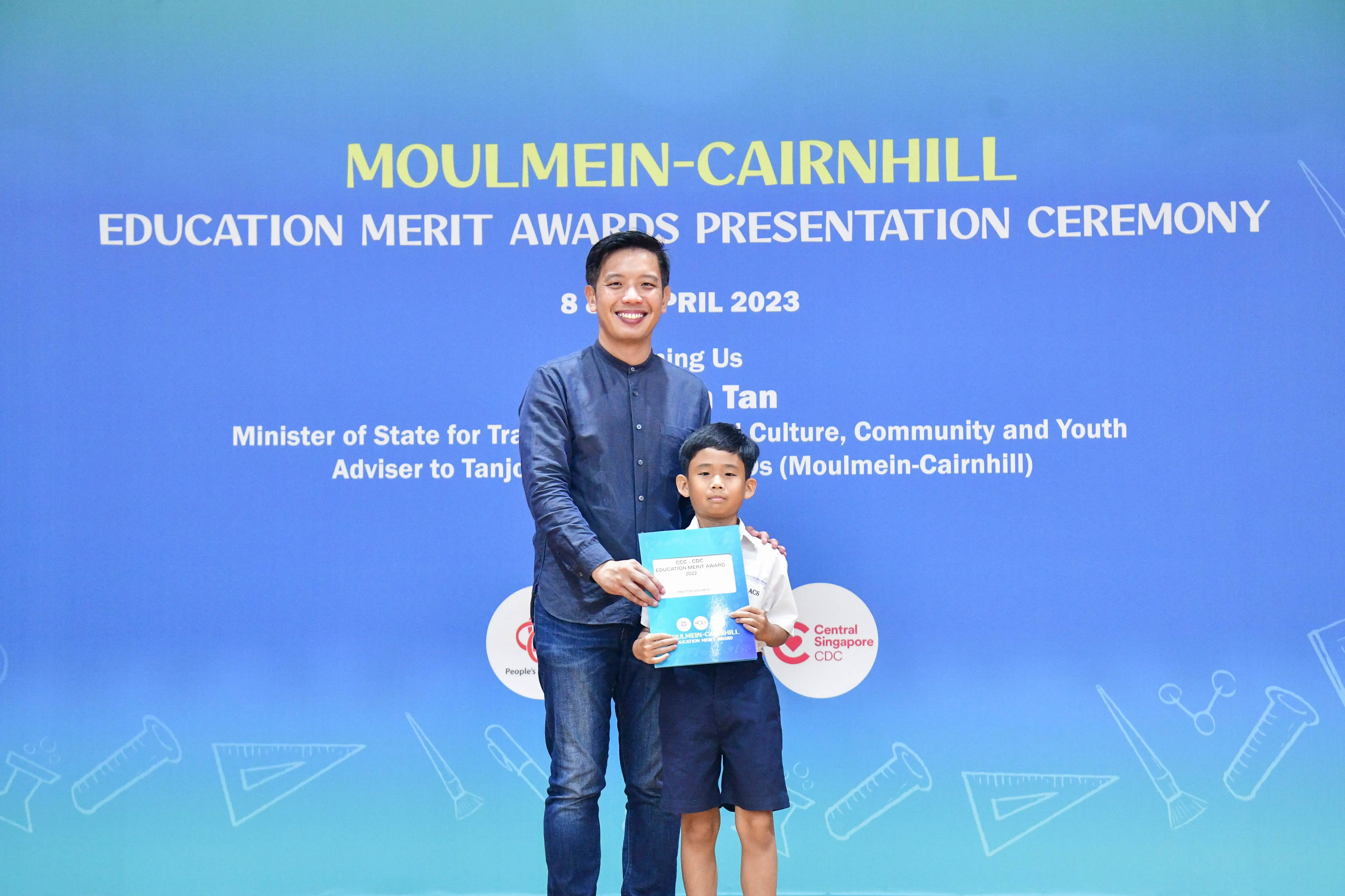A man and a young boy hold a certificate in front of a blue Moulmein-Cairnhill Education Merit Awards banner.