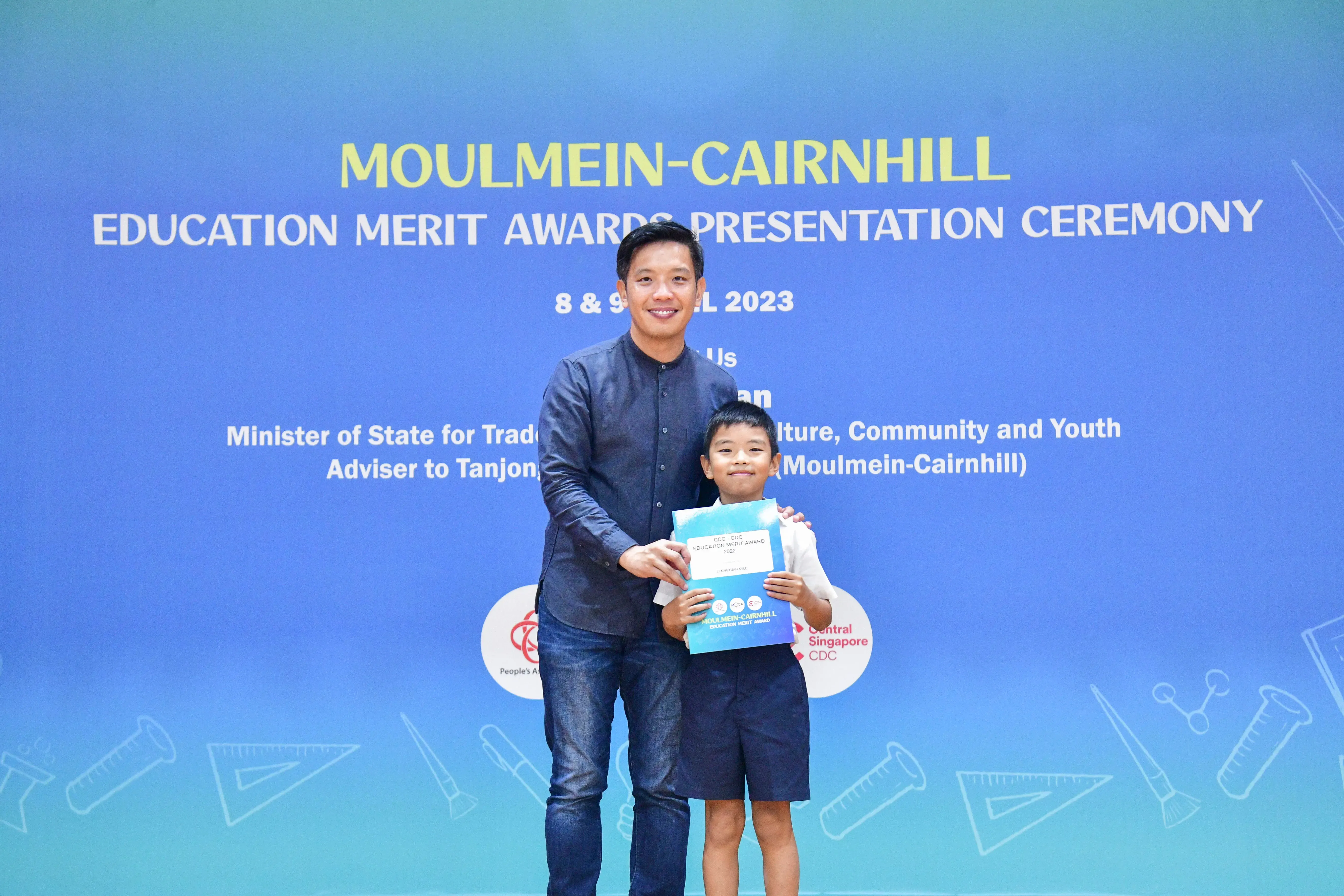 A man and a young boy hold a certificate in front of a Moulmein-Cairnhill Education Merit Awards banner.