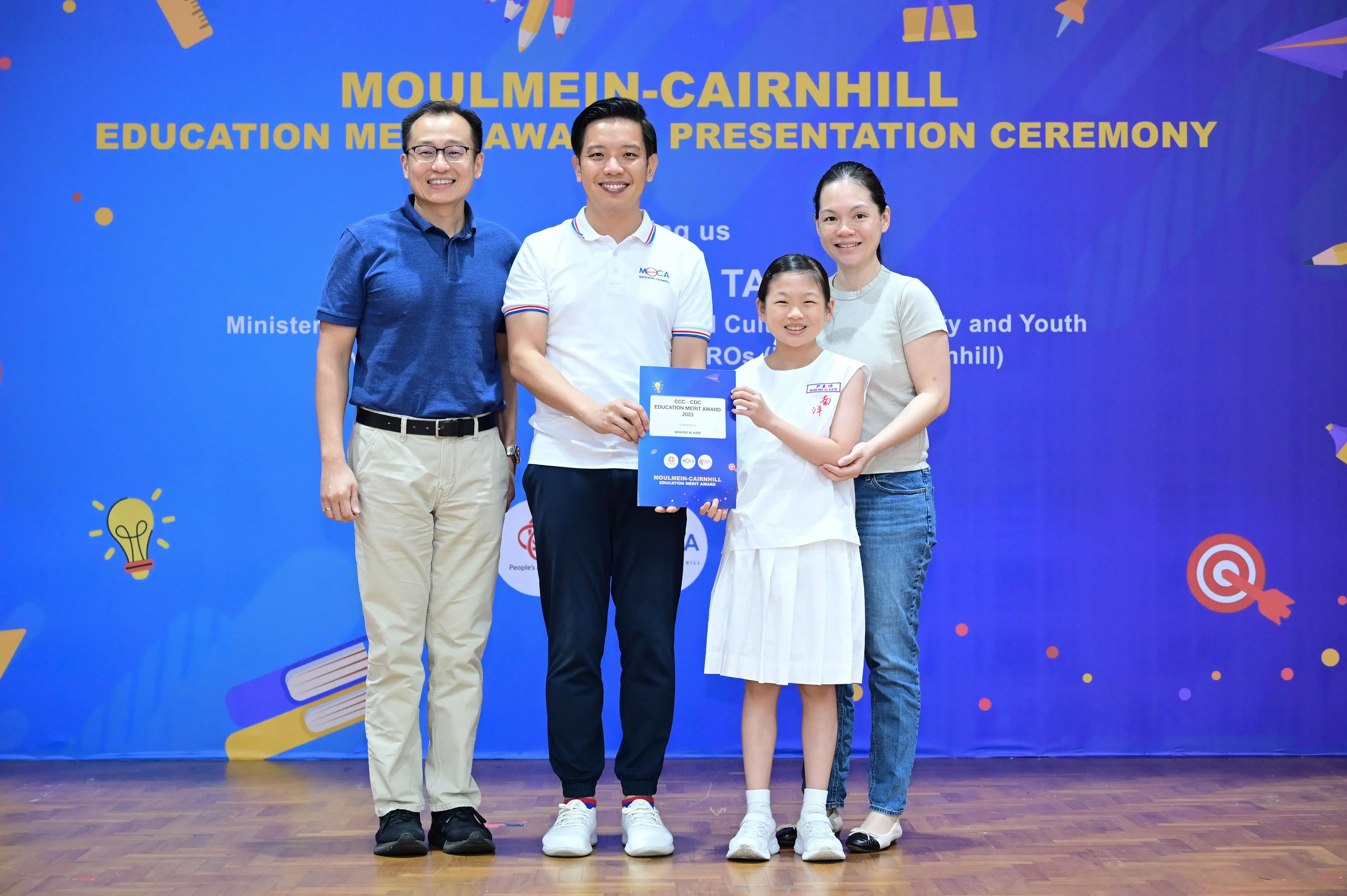 Four people pose at the Moulmein-Cairnhill Education Merit Award Presentation Ceremony, with a girl holding a blue certificate.