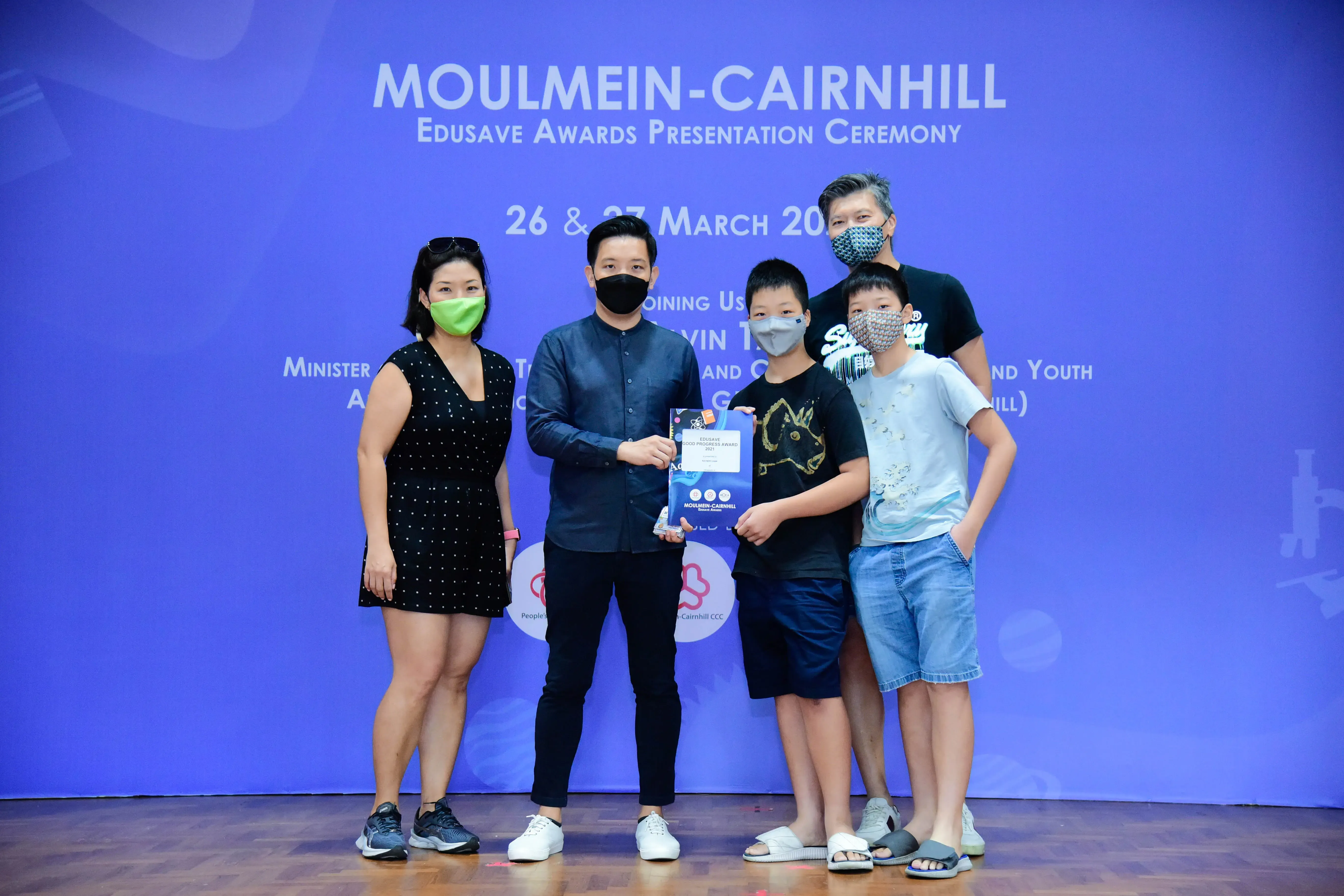 Five masked people pose at the Moulmein-Cairnhill Edusave Awards Presentation Ceremony, with a boy holding an award.