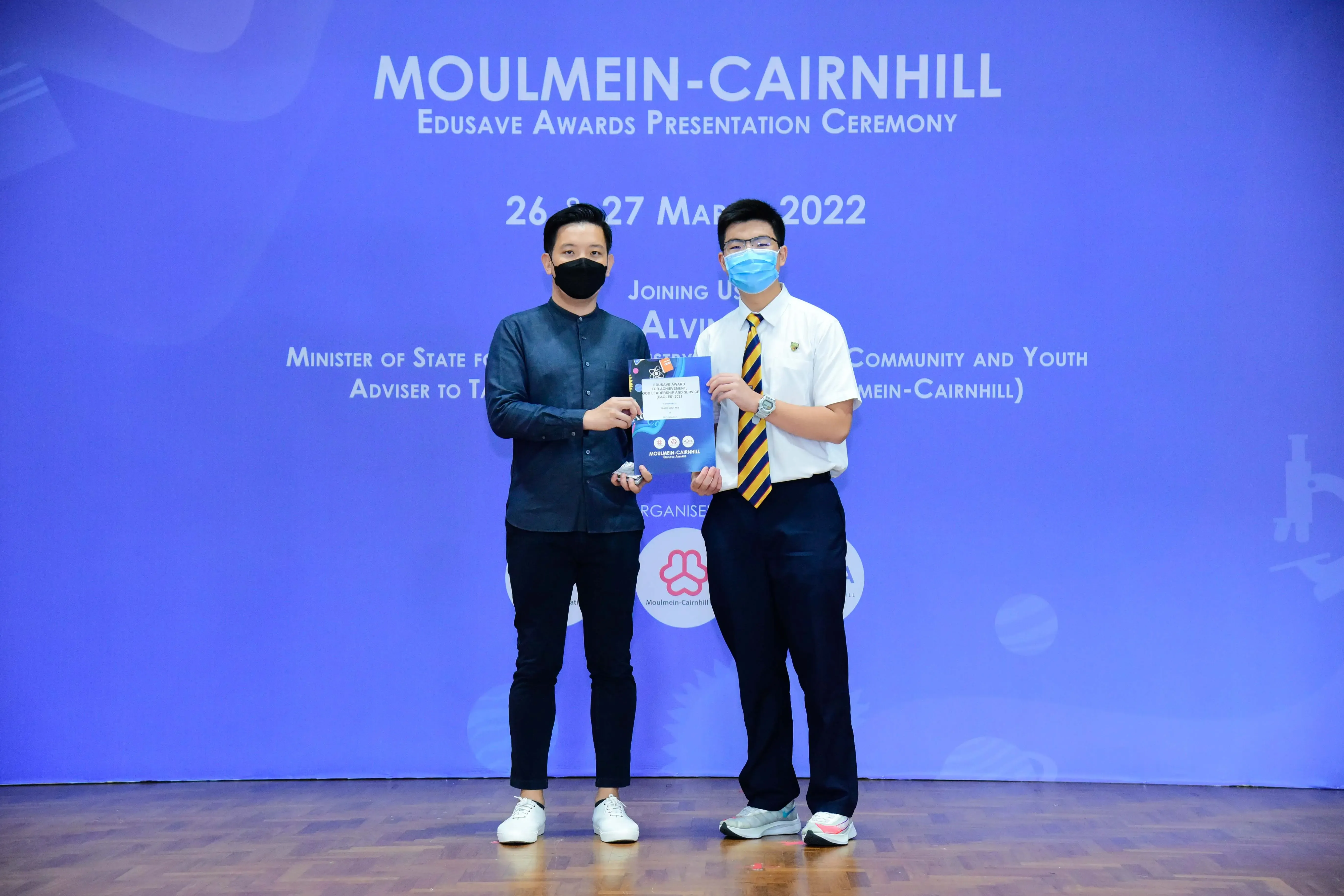 Two masked men hold an award at the Moulmein-Cairnhill Edusave Awards Presentation Ceremony, March 2022.