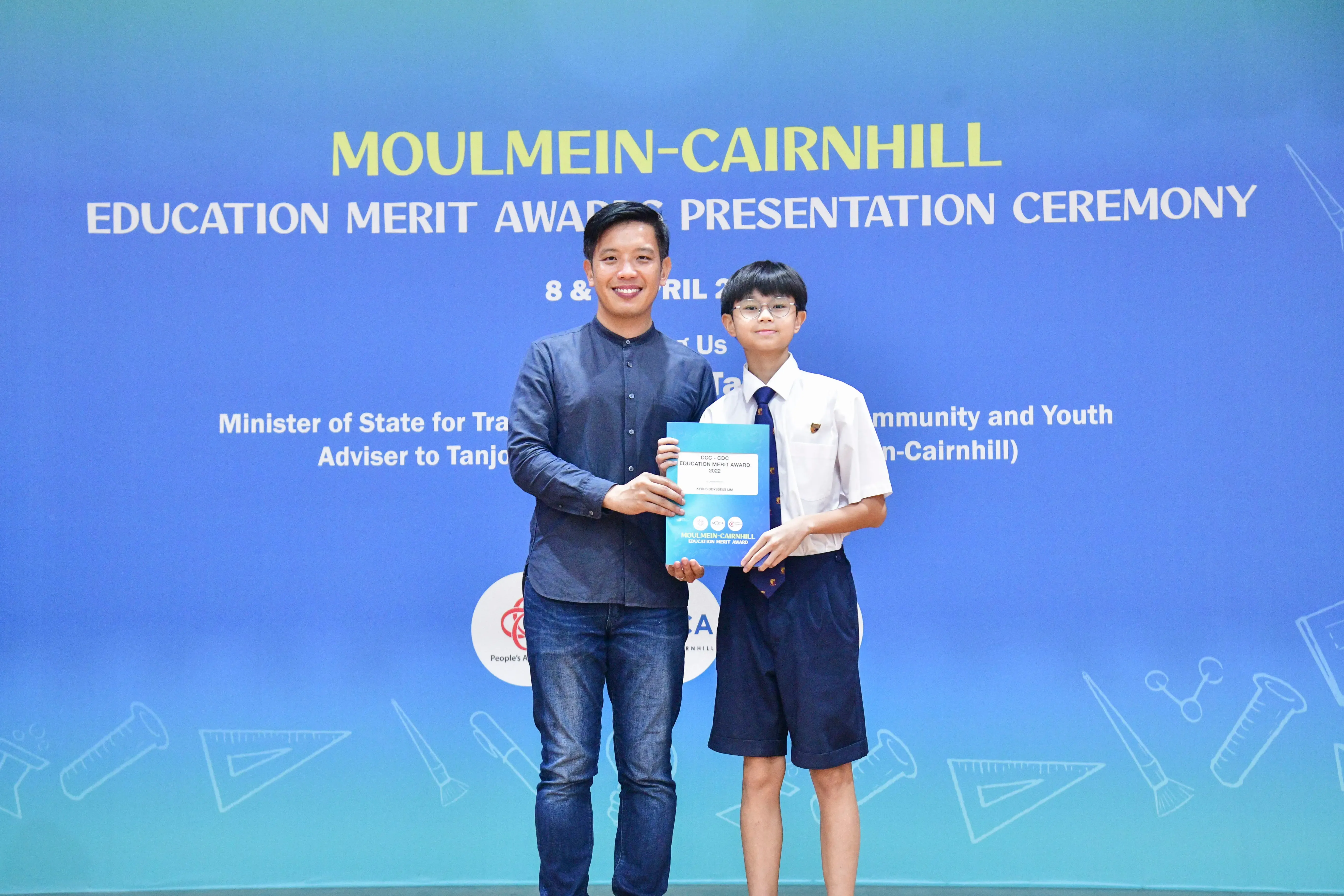An adult and a young student hold a certificate together at the Moulmein-Cairnhill Education Merit Award ceremony.