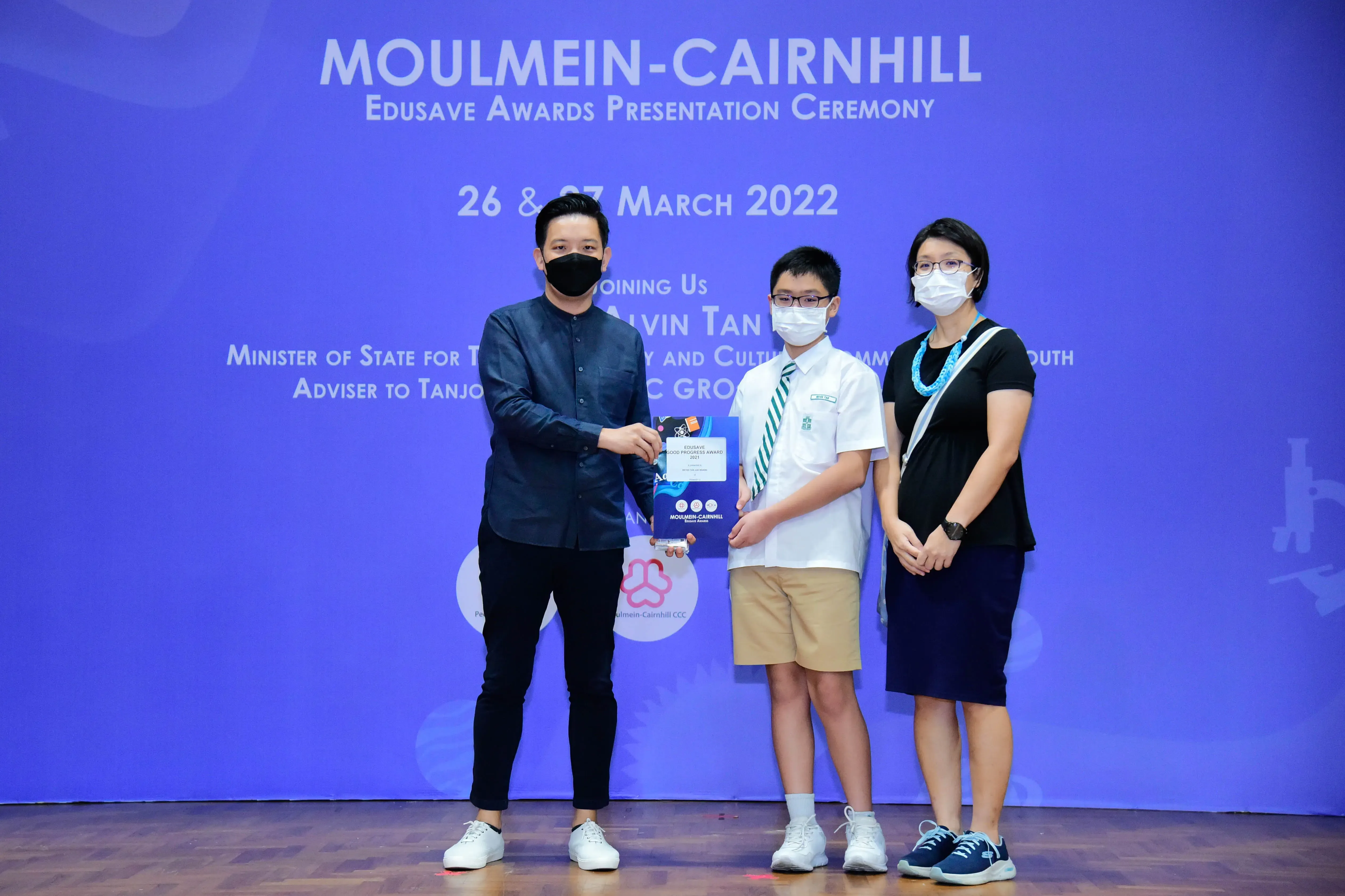 Three masked people stand on stage at the Moulmein-Cairnhill Edusave Awards Ceremony, with an adult handing a certificate to a student.