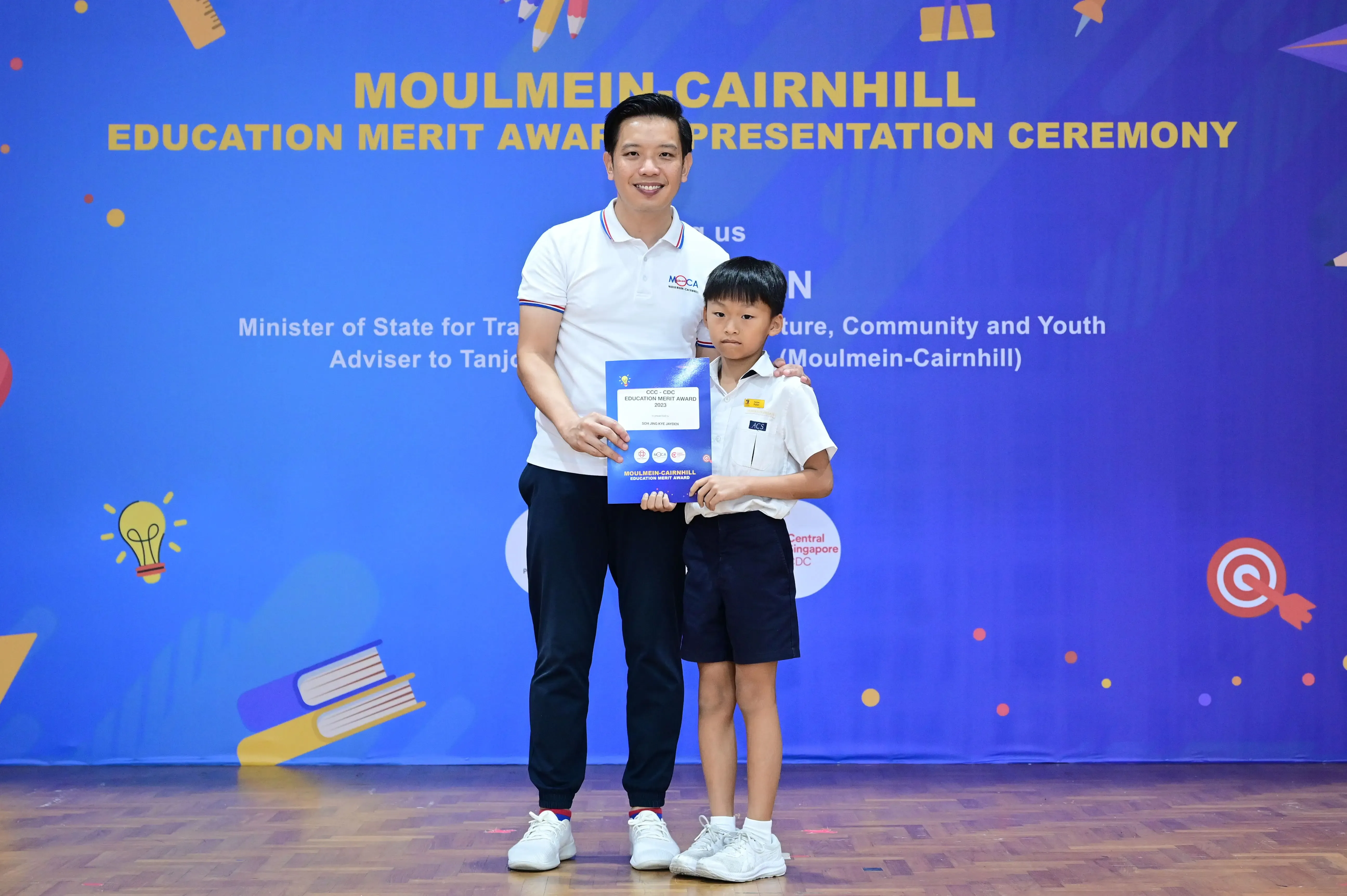 A man and a young boy hold a blue award certificate in front of a Moulmein-Cairnhill Education Merit Award banner.