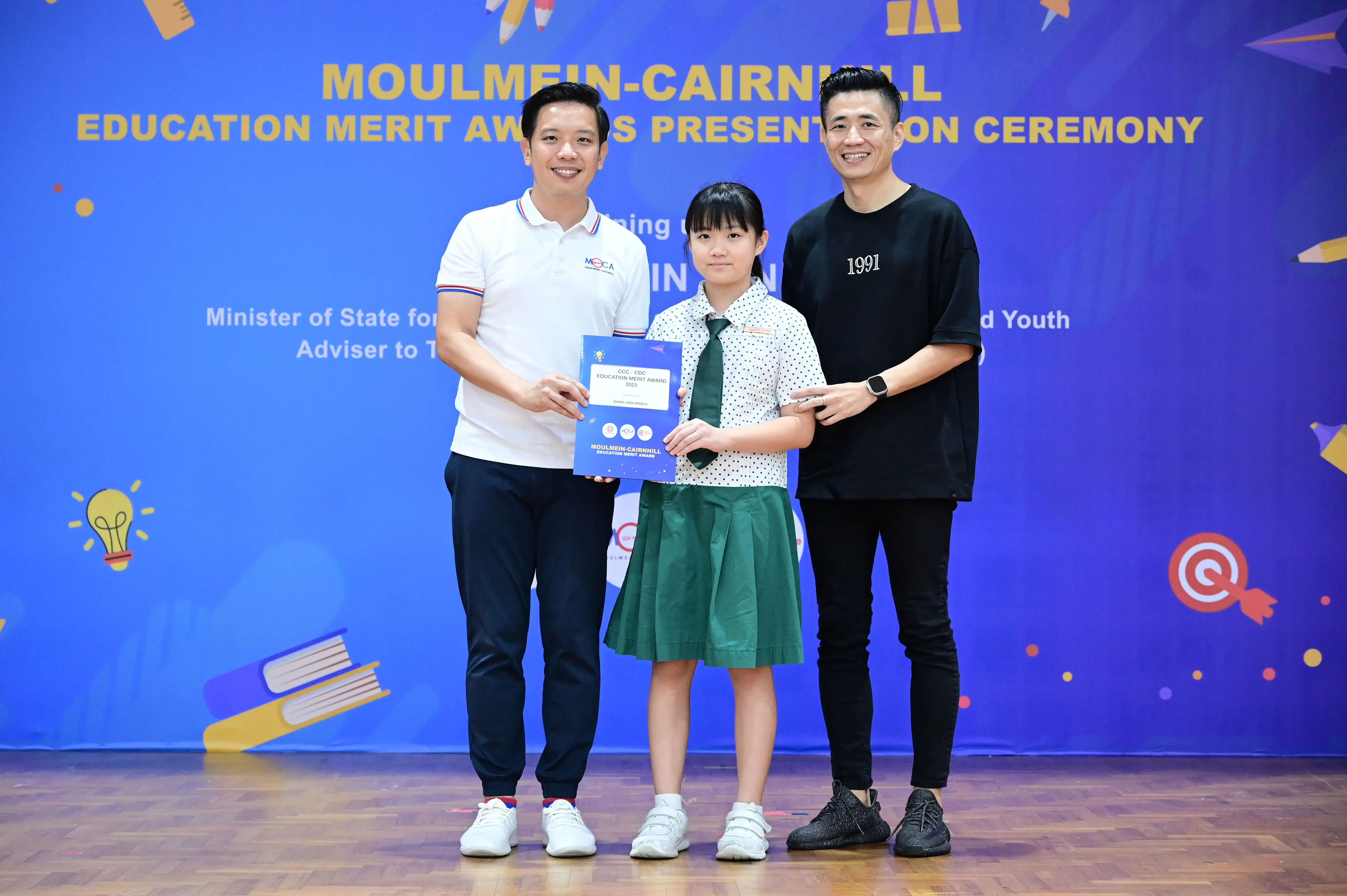 Two men and a girl in school uniform hold a certificate at the Moulmein-Cairnhill Education Merit Awards ceremony.