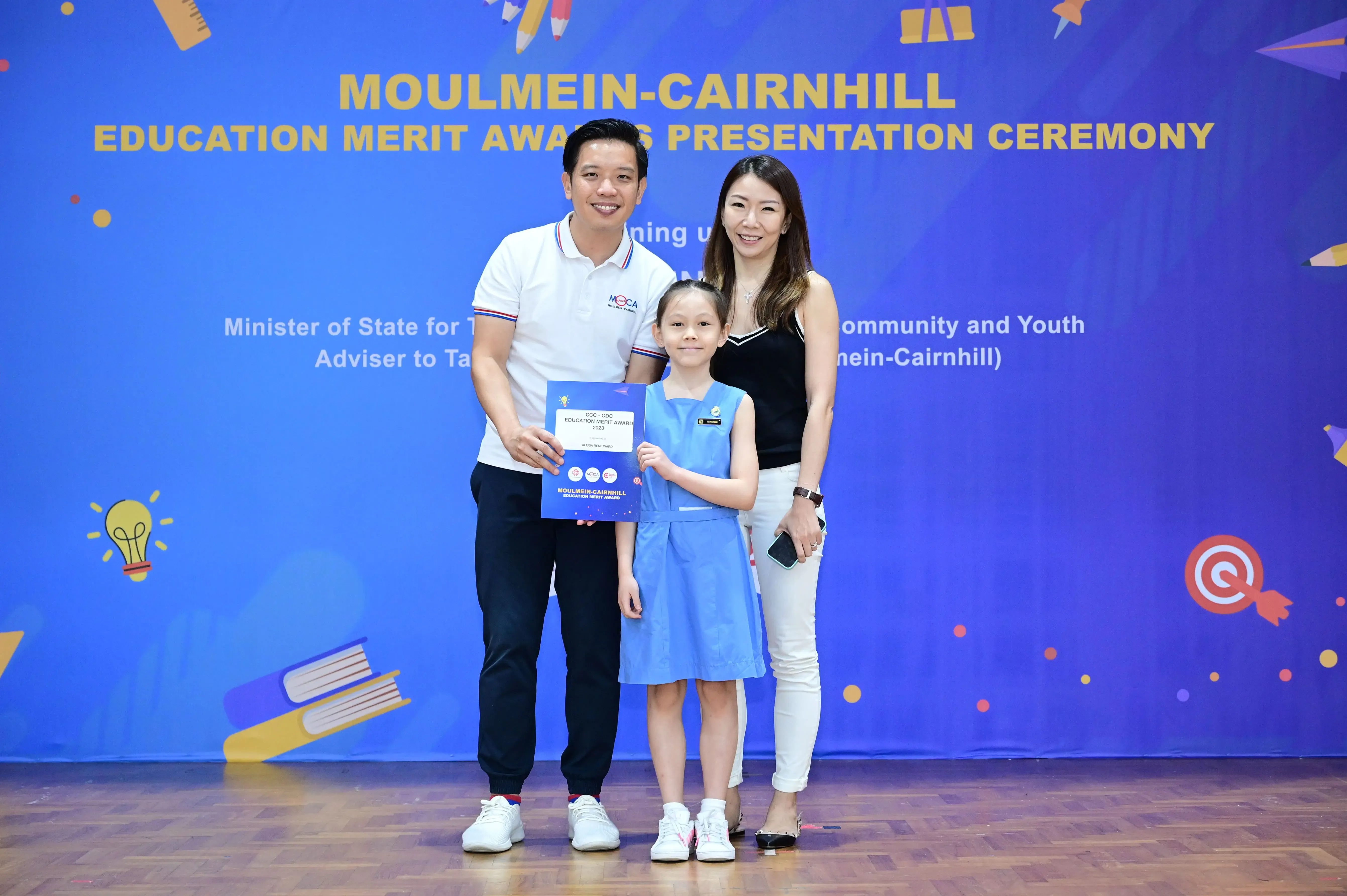 A girl in a blue school uniform holds an award certificate, flanked by two smiling adults, at the Moulmein-Cairnhill Education Merit Awards ceremony.