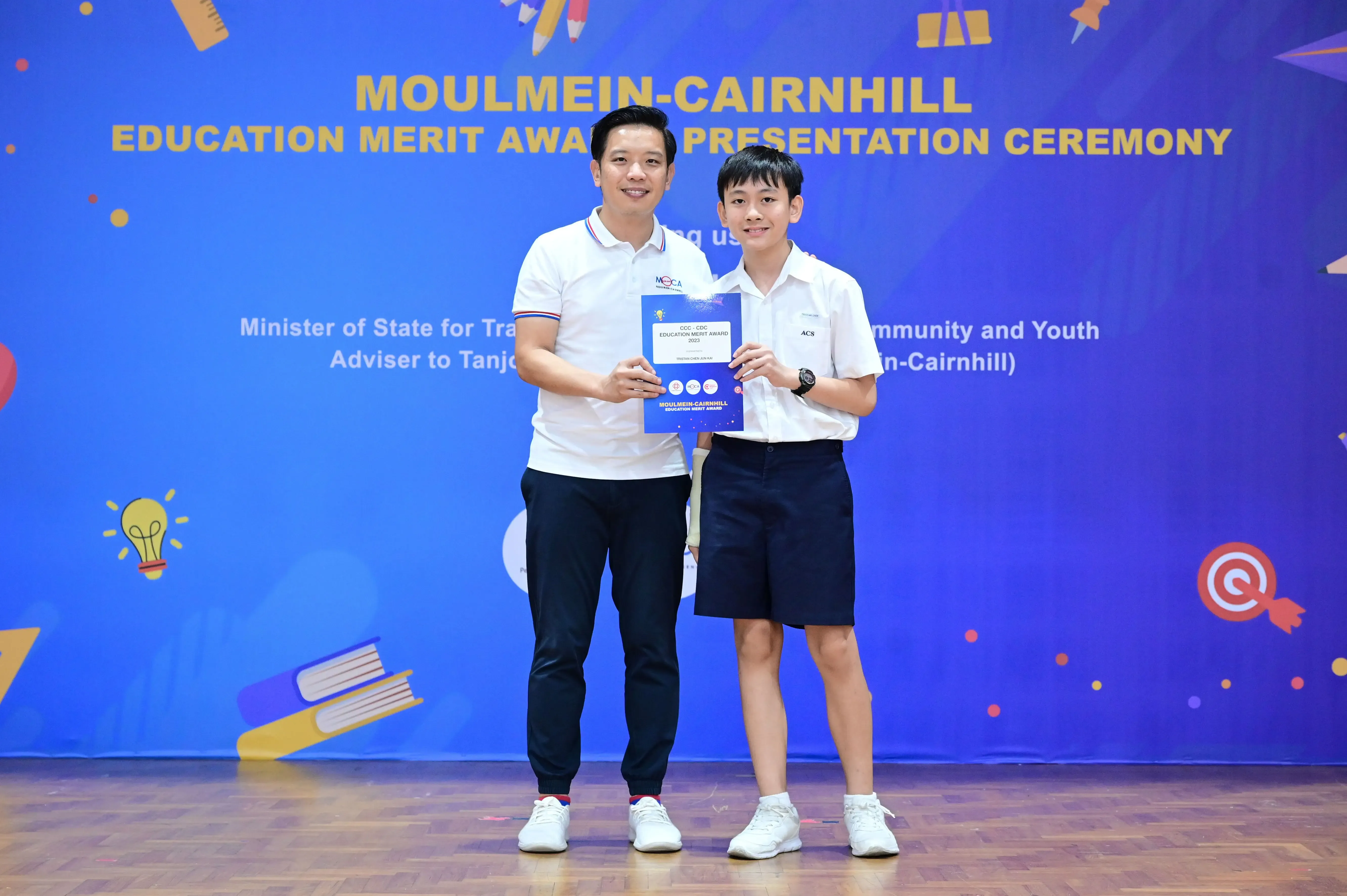 An adult and a young student hold an award certificate at the Moulmein-Cairnhill Education Merit Award Presentation Ceremony.