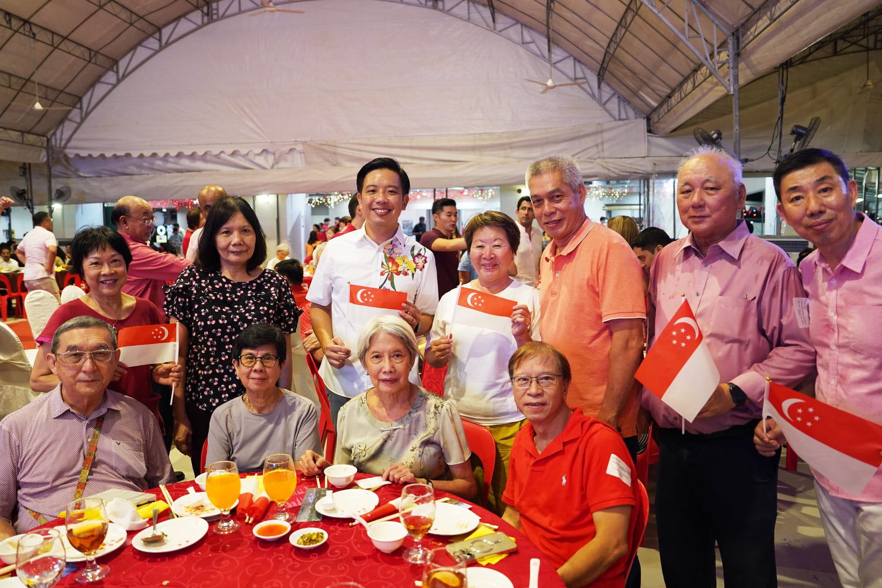 A group of smiling people at a banquet holding small Singapore flags, seated and standing around a red dining table.