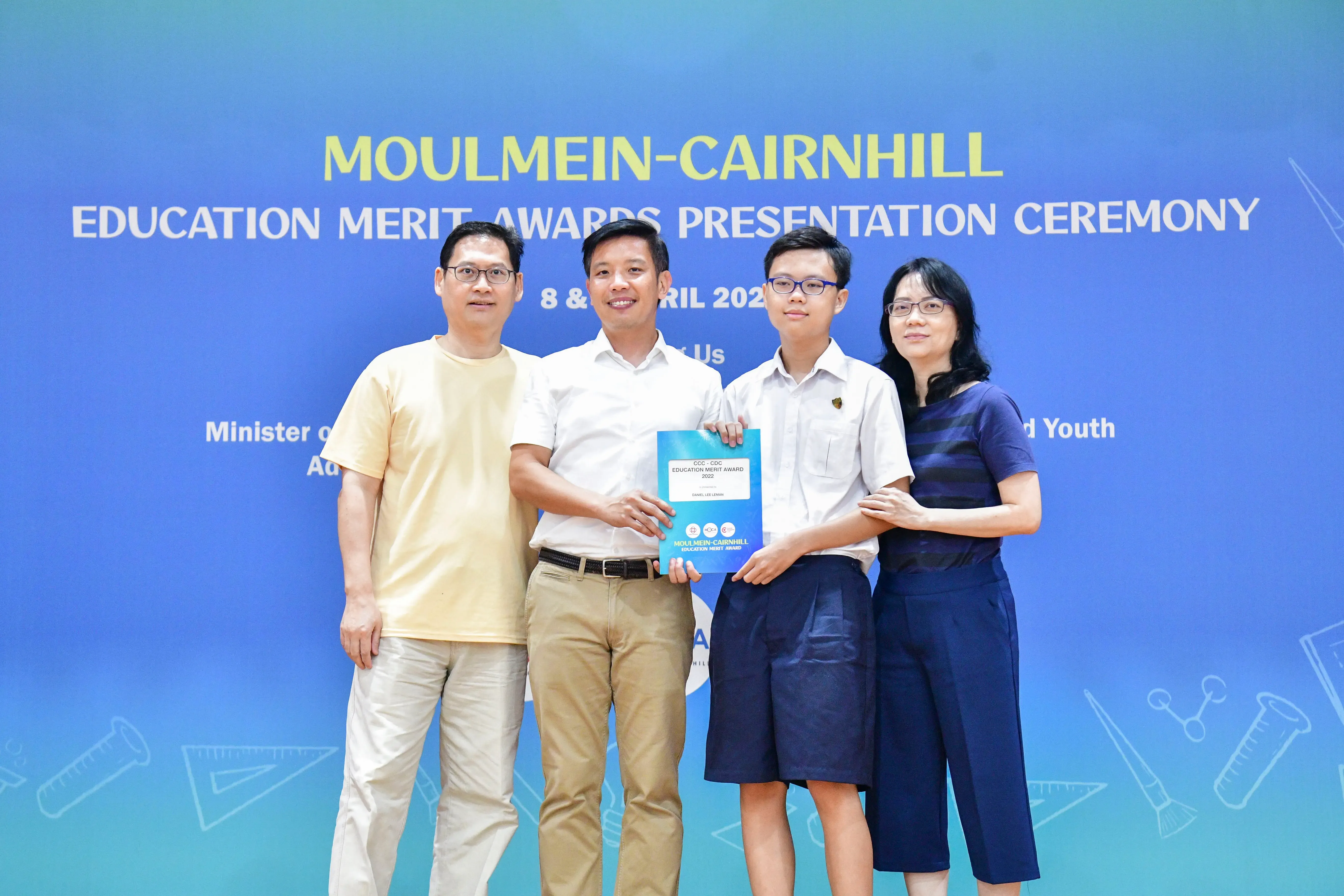 Four people pose in front of a blue Moulmein-Cairnhill Education Merit Awards Presentation Ceremony banner.
