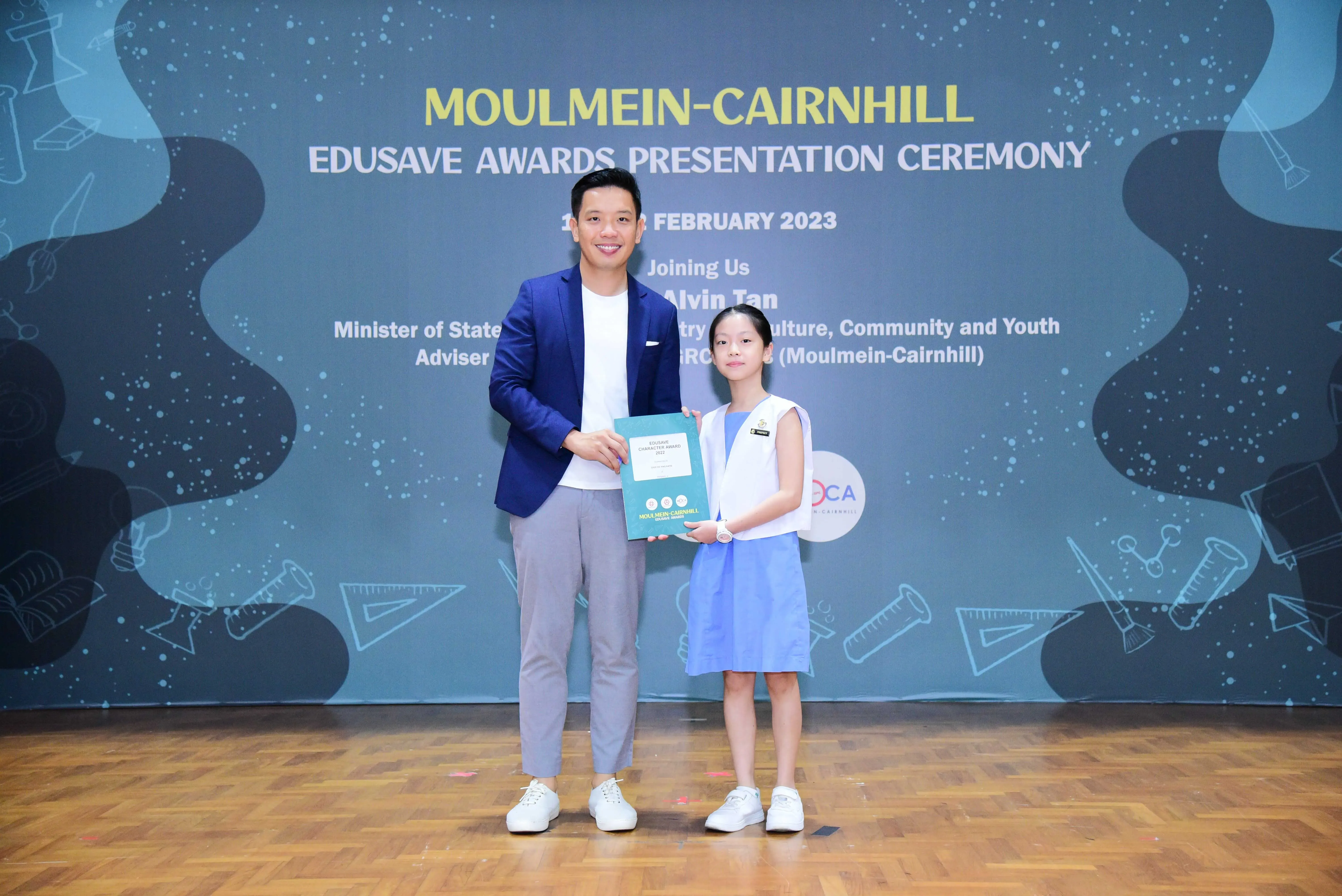 A man in a blue blazer and a young girl in a blue dress hold a certificate at the Moulmein-Cairnhill Edusave Awards 2023.