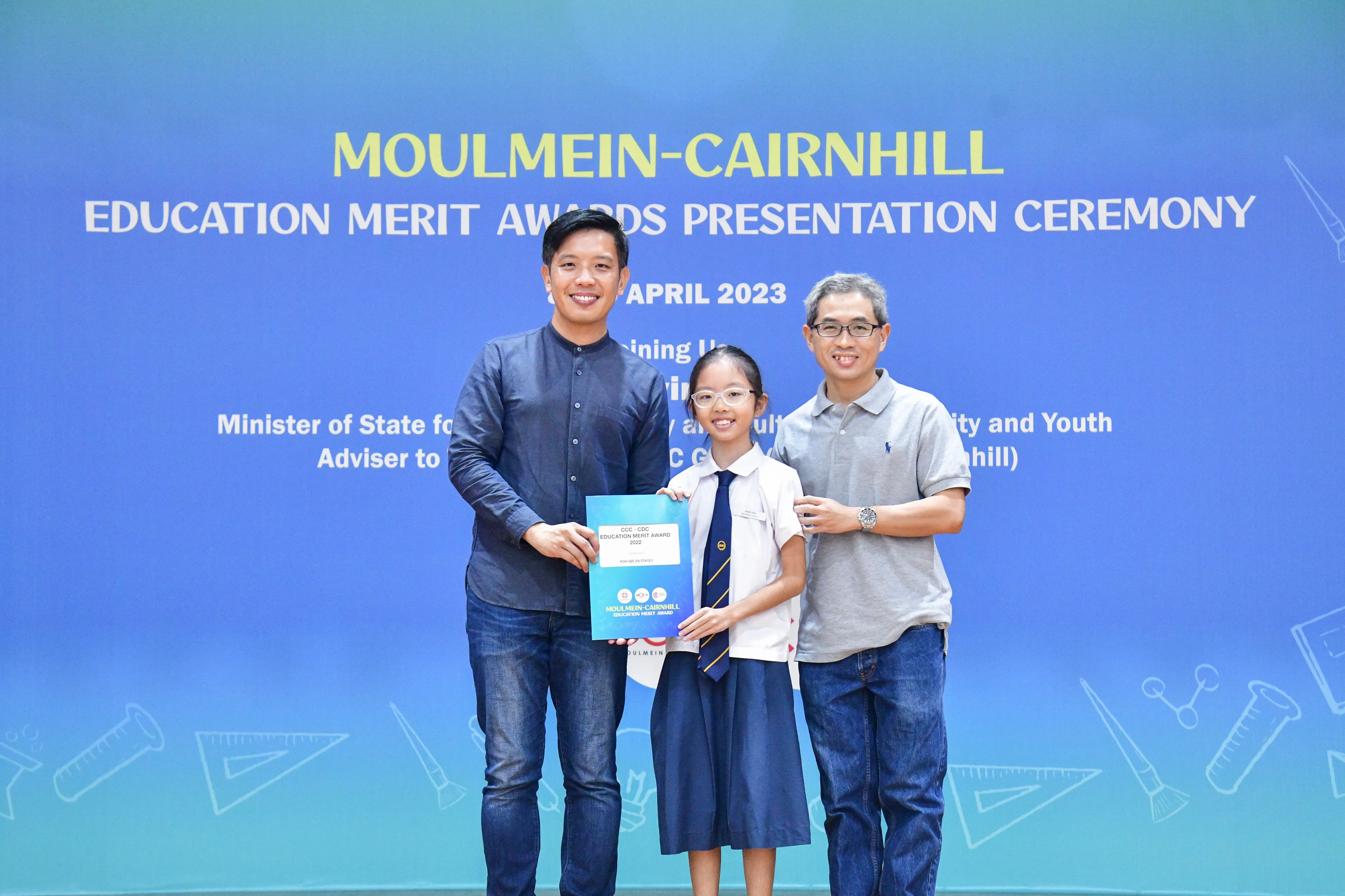 Two men and a girl in school uniform hold a certificate at the Moulmein-Cairnhill Education Merit Awards April 2023.
