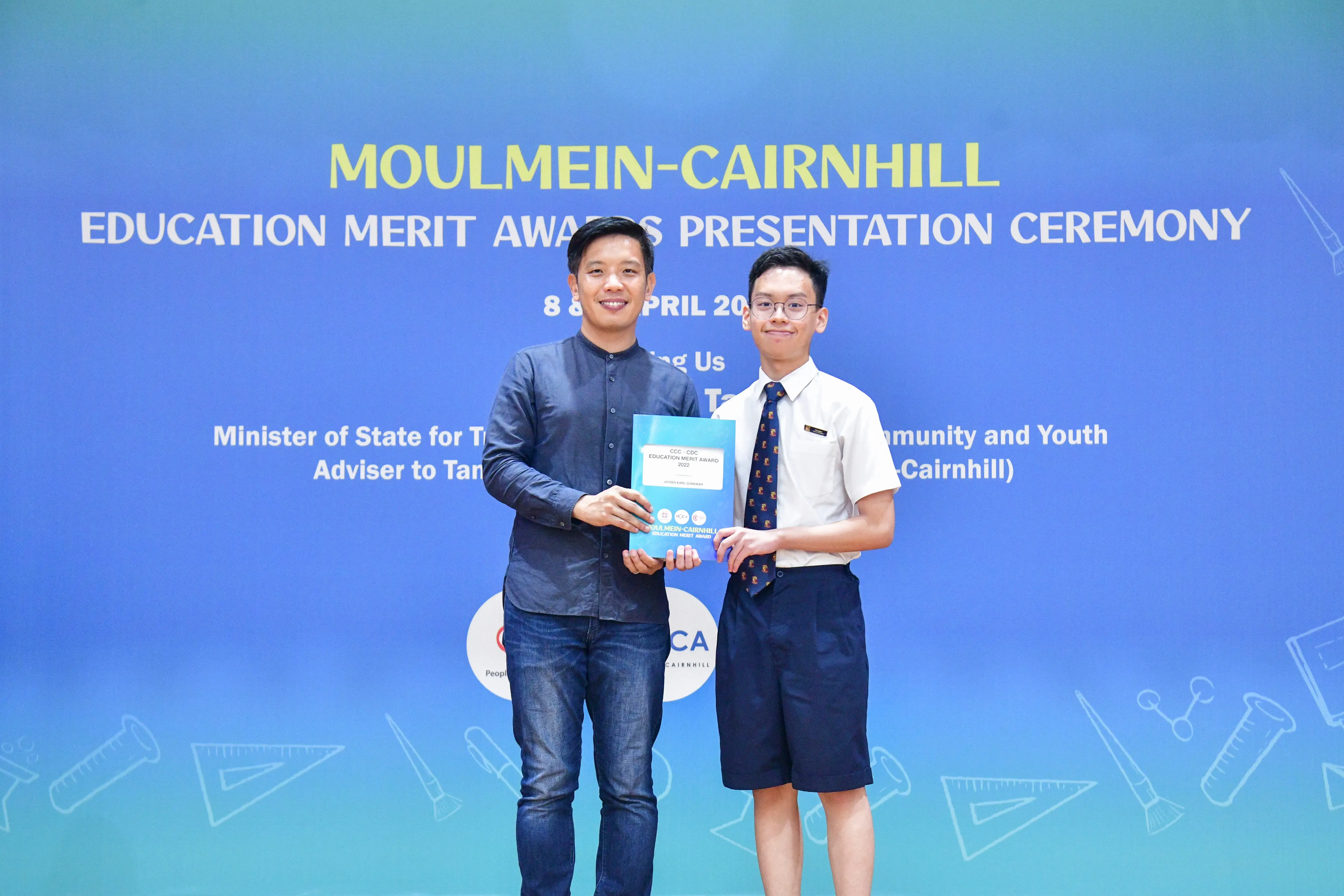 Two people hold a certificate together in front of a Moulmein-Cairnhill Education Merit Awards Presentation Ceremony banner.