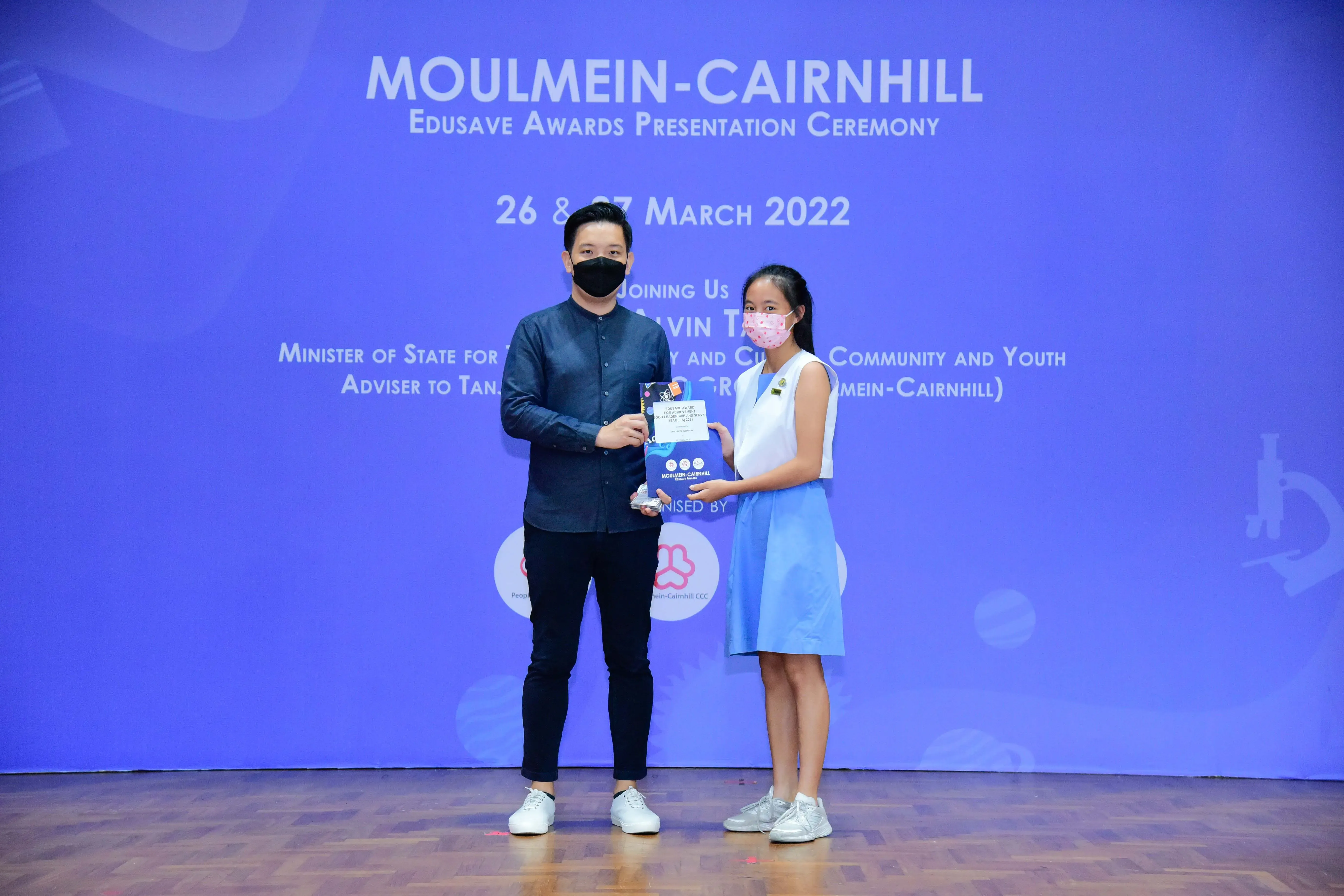 A masked man presents an award to a masked girl in a blue dress at the Moulmein-Cairnhill Edusave Awards Ceremony 2022.