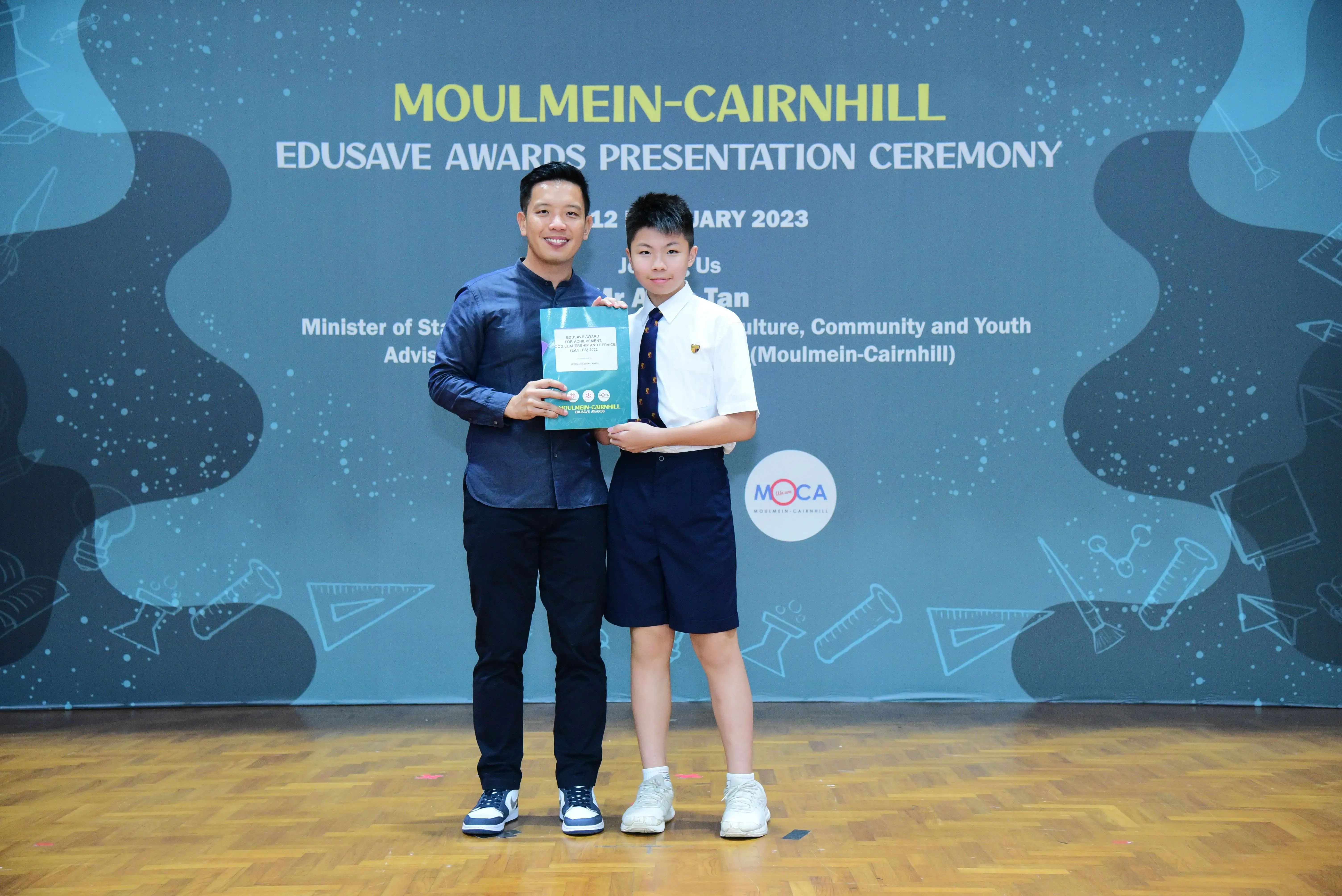 Two people hold a certificate together on stage at the Moulmein-Cairnhill Edusave Awards Presentation Ceremony 2023.