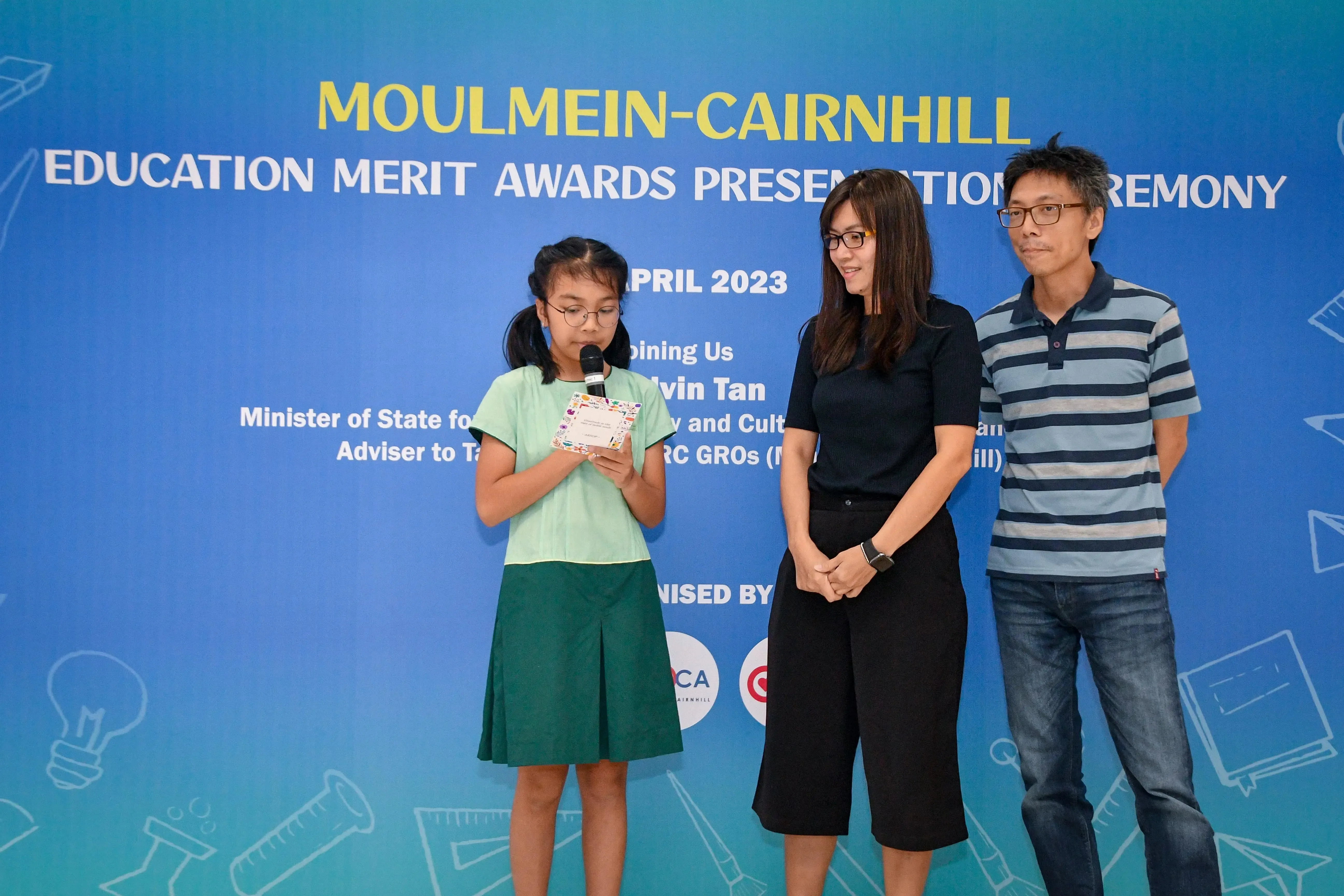A girl speaks into a microphone at the Moulmein-Cairnhill Education Merit Awards Ceremony, April 2023, with two adults beside her.
