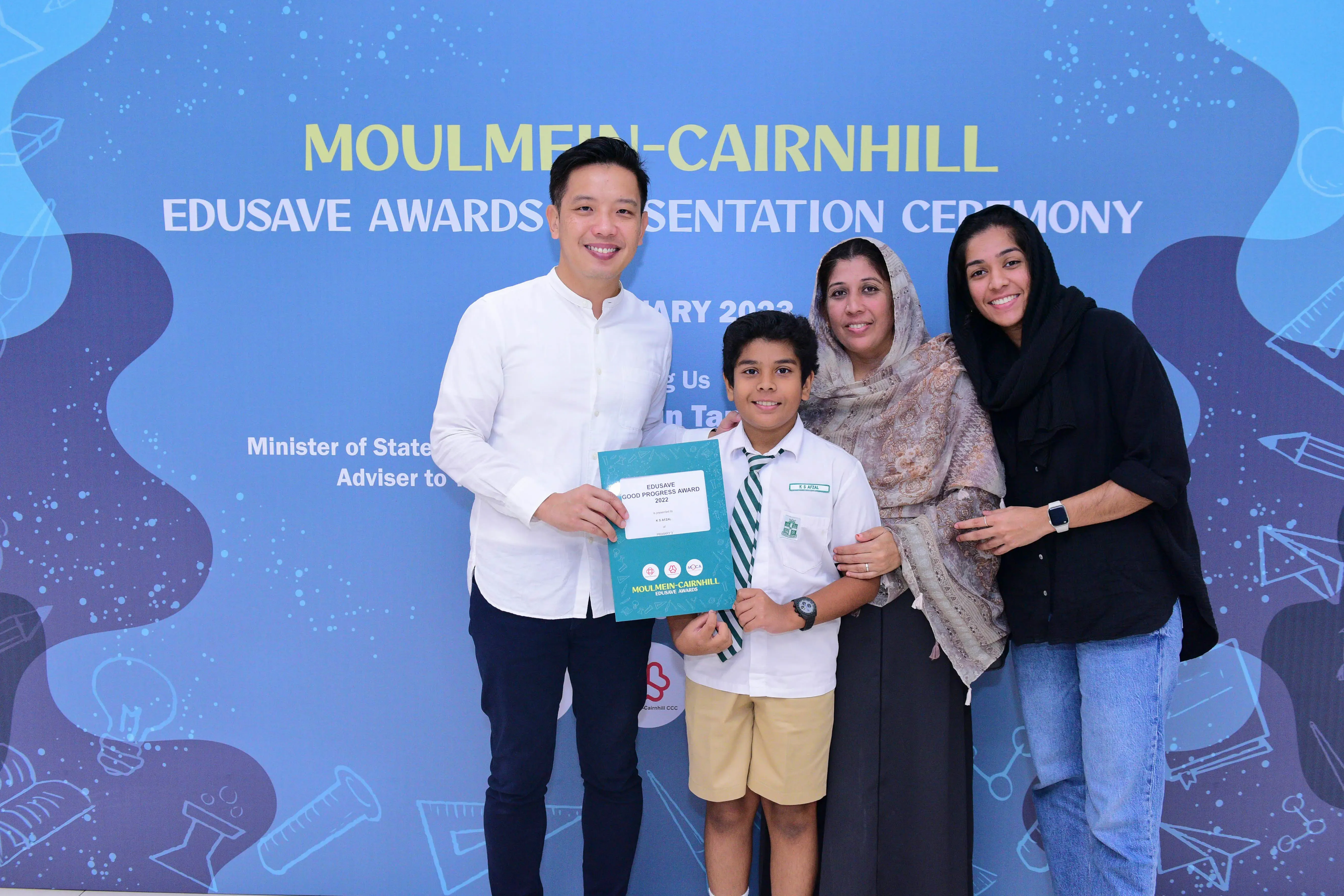 A boy in school uniform holds an Edusave award certificate, posing with three smiling adults at an awards ceremony.