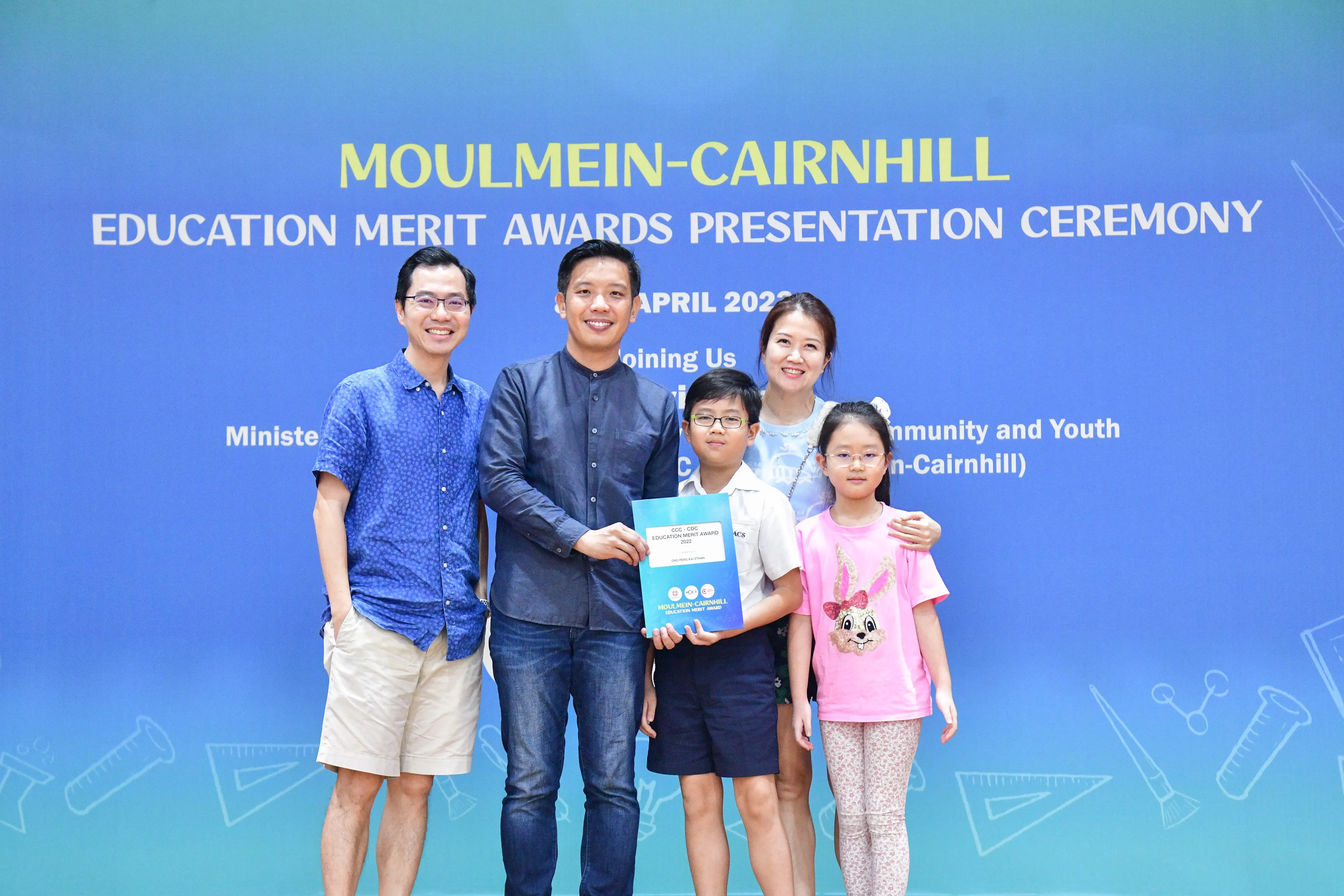 Five people pose before a Moulmein-Cairnhill Education Merit Awards banner, with a boy holding a certificate.