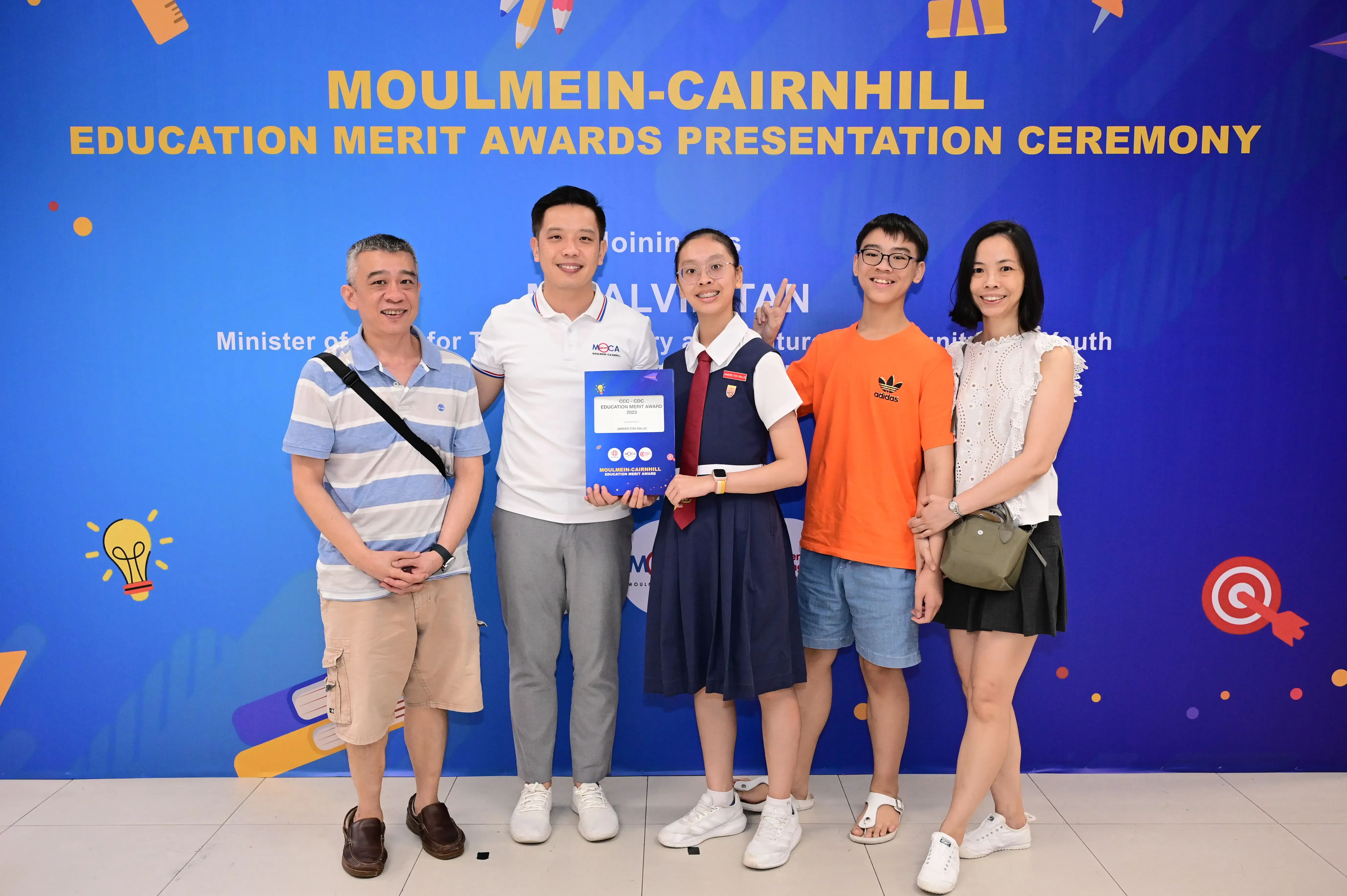 Five people pose before a Moulmein-Cairnhill Education Merit Awards banner, with a girl in uniform holding a certificate.