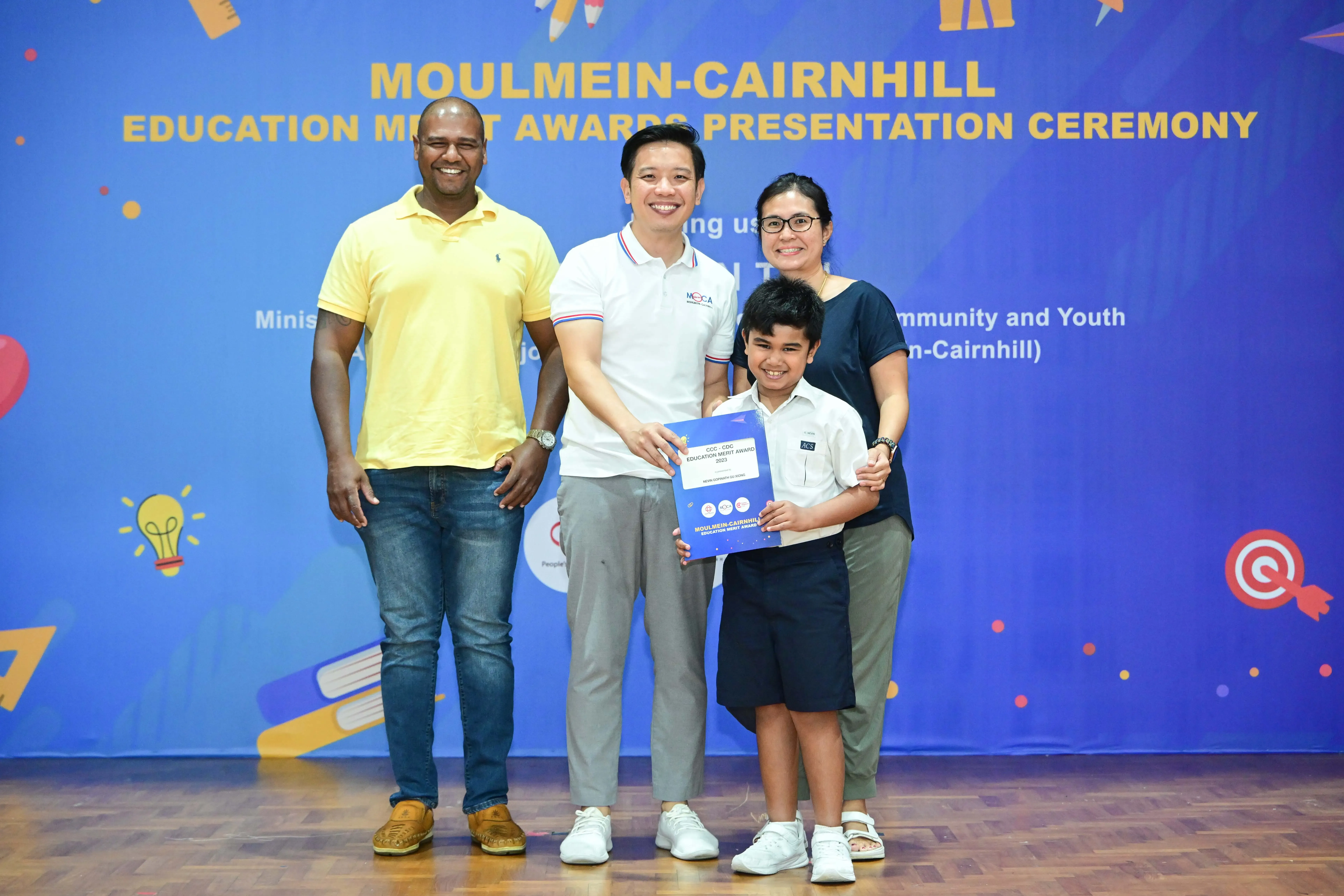 Four people pose at the Moulmein-Cairnhill Education Merit Awards Presentation Ceremony, with a boy holding a certificate.