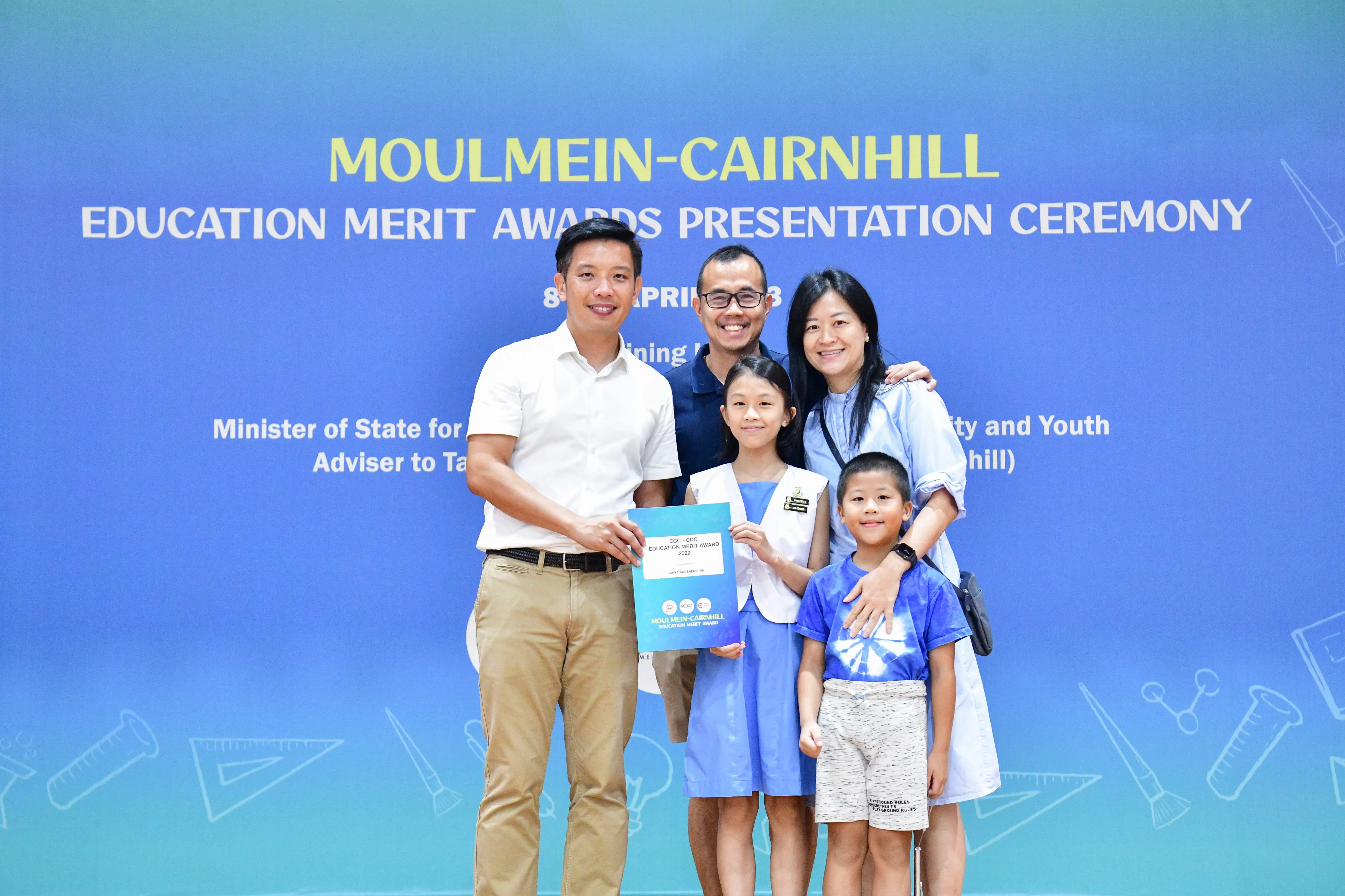 Five people pose in front of a blue Moulmein-Cairnhill Education Merit Awards Presentation Ceremony banner.