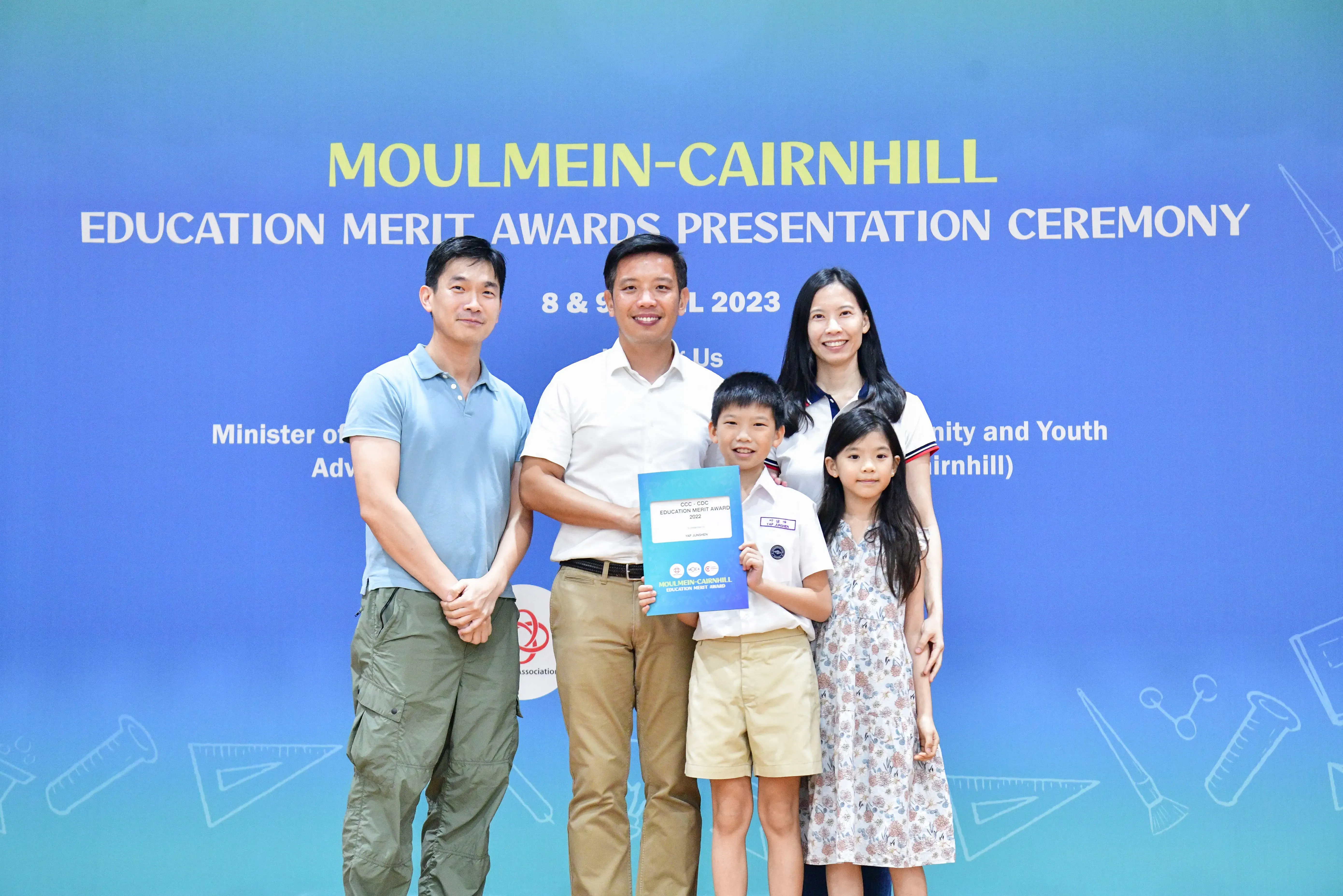 Five people pose before a blue banner for the Moulmein-Cairnhill Education Merit Awards 2023, with a boy holding a certificate.