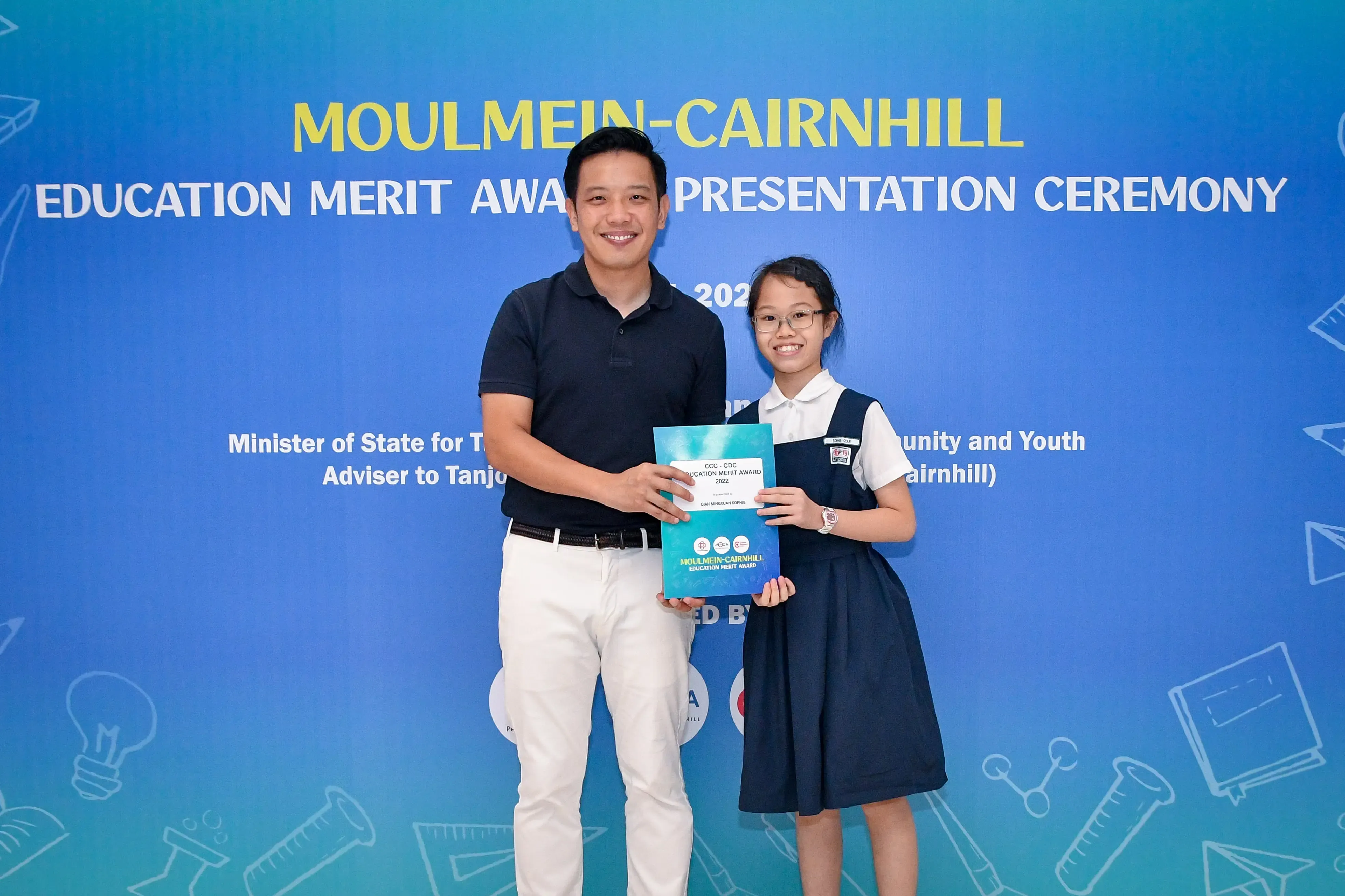 A man and a young girl in school uniform hold an award certificate at the Moulmein-Cairnhill Education Merit Award ceremony.