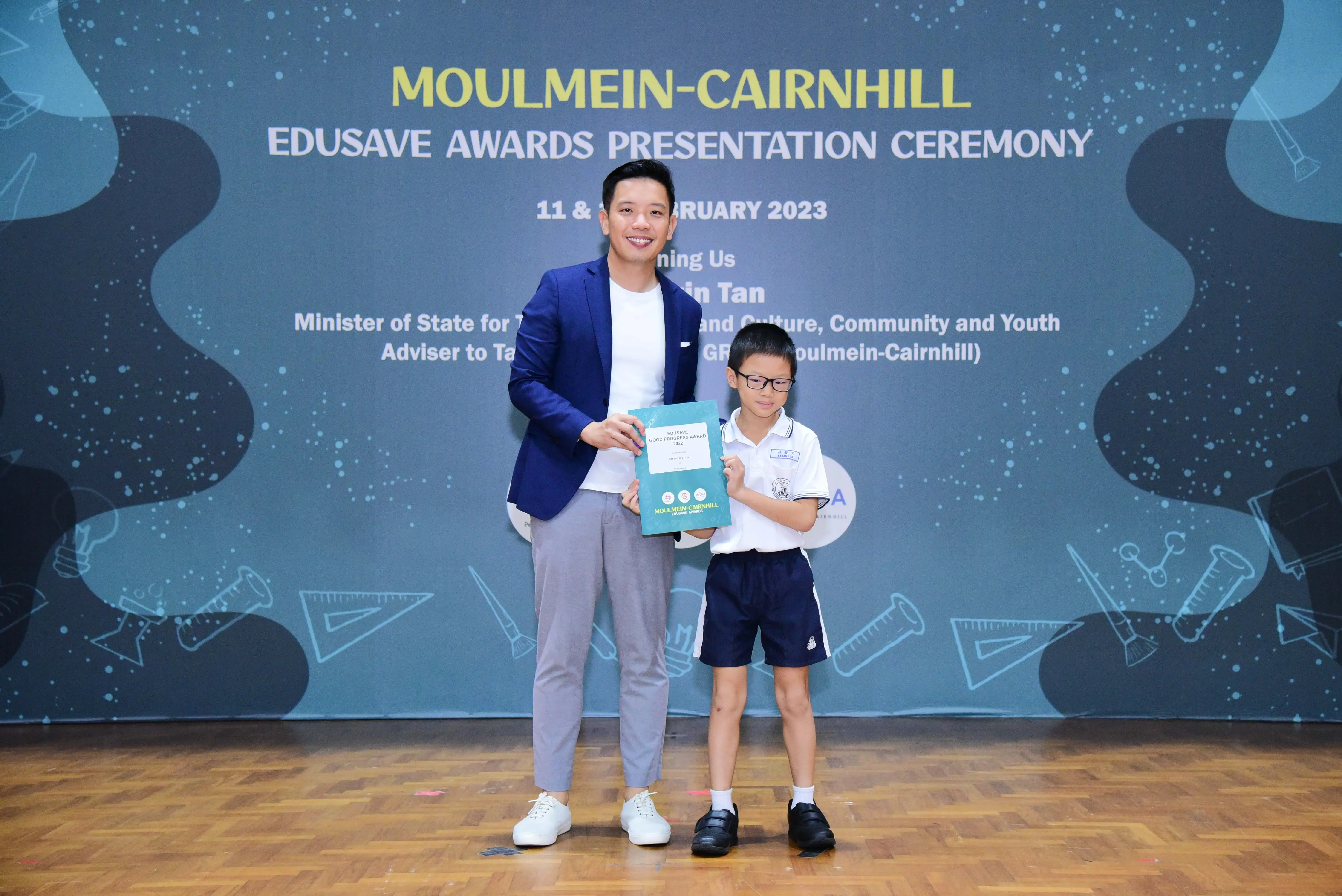 A man in a blue blazer and a young student hold an award at the Moulmein-Cairnhill Edusave Awards Ceremony 2023.
