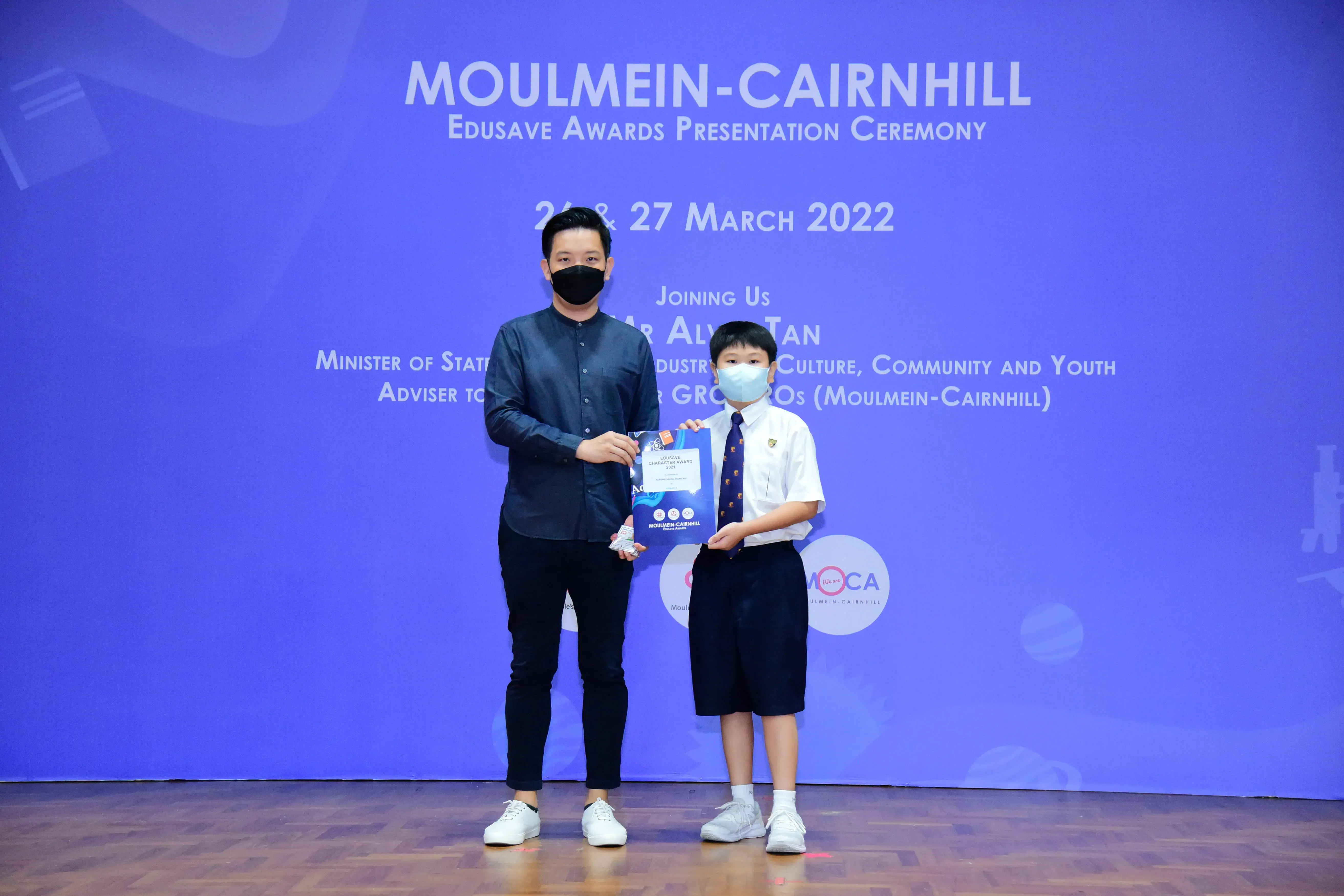 An adult and a young student in uniform hold a certificate at the Moulmein-Cairnhill Edusave Awards Ceremony, March 2022.