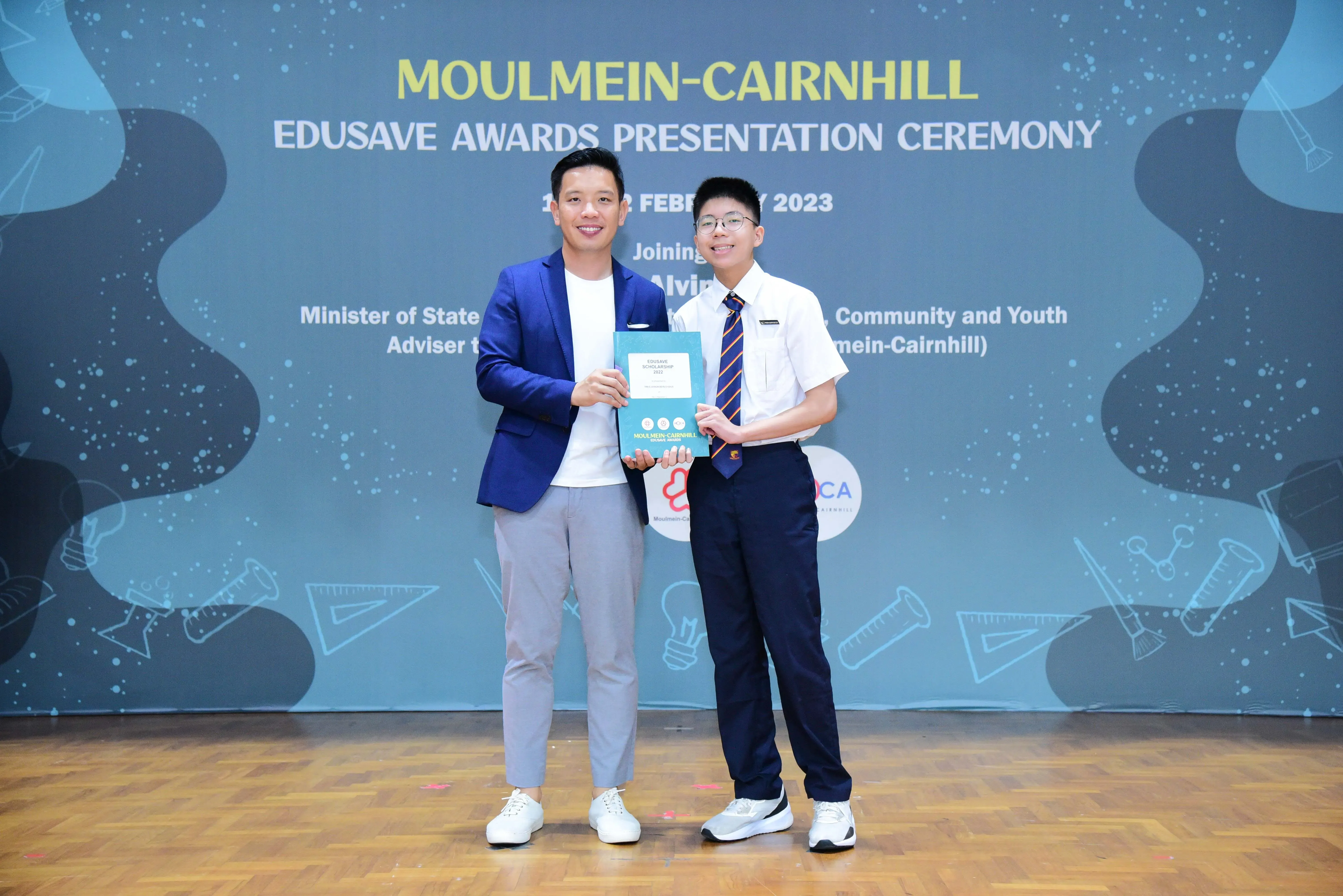 Two people hold a certificate together on stage at the Moulmein-Cairnhill Edusave Awards Presentation Ceremony 2023.