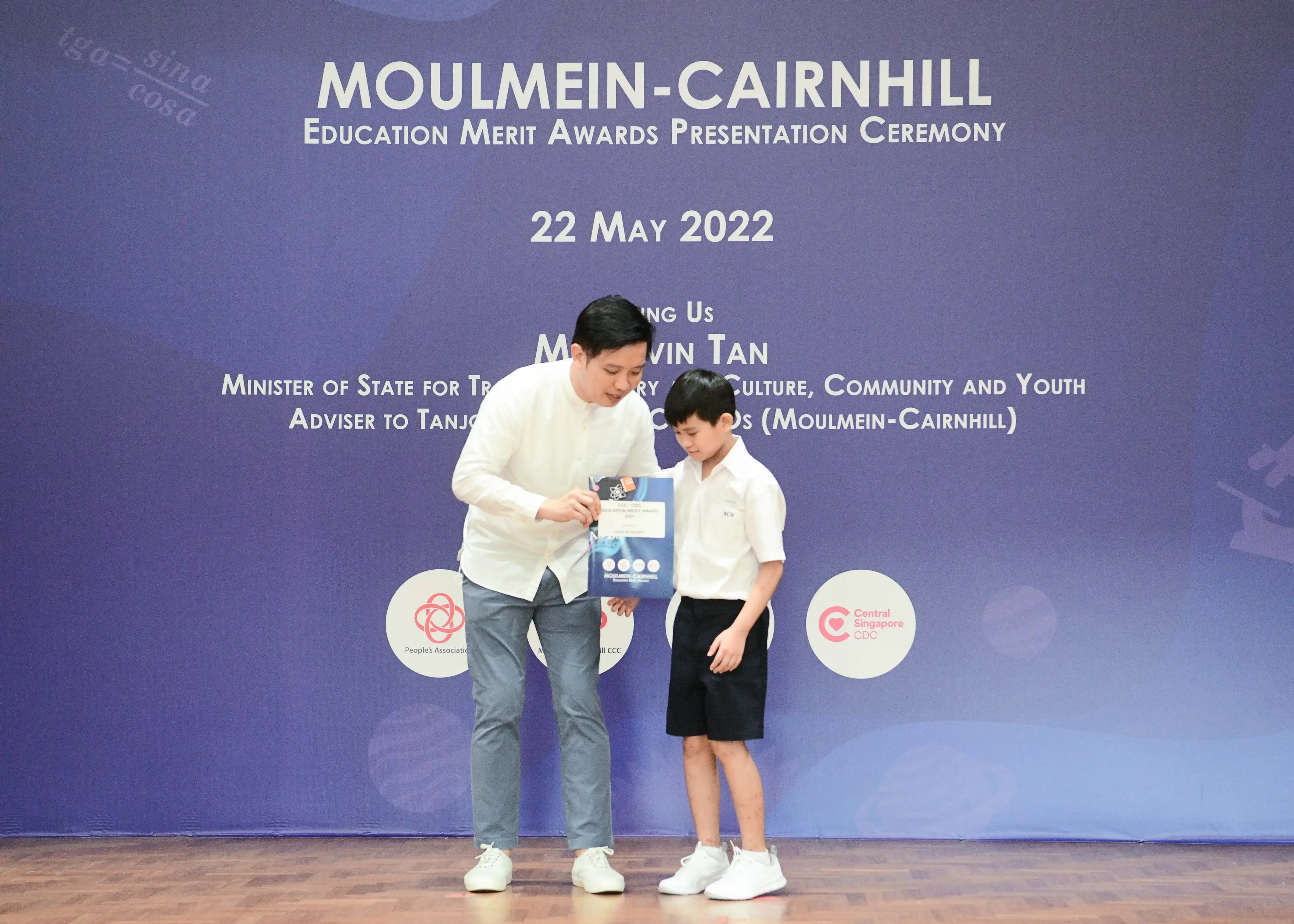 An adult in white shirt hands a booklet to a young boy at the Moulmein-Cairnhill Education Merit Awards ceremony on 22 May 2022.