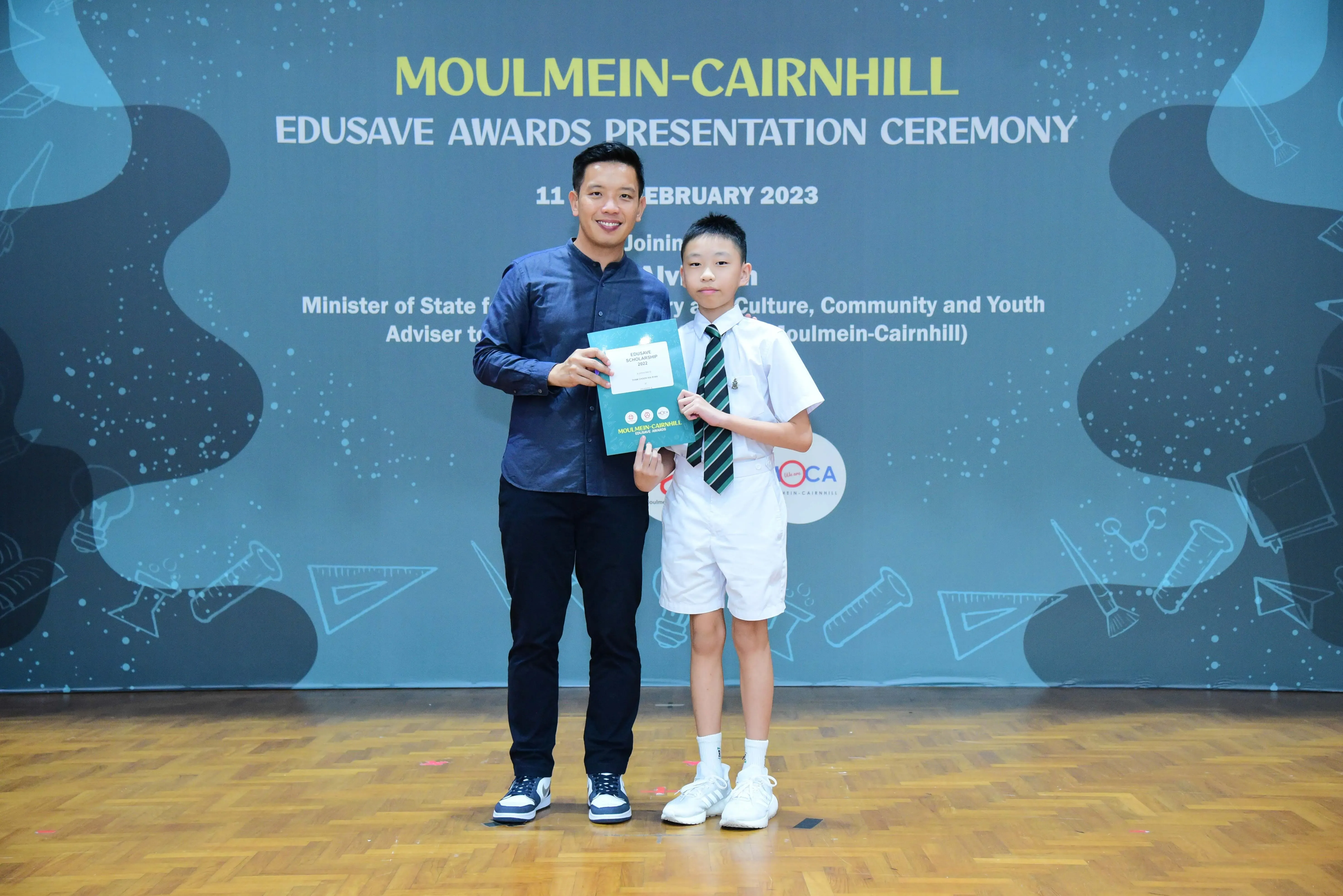 An adult and a uniformed student hold a certificate at the Moulmein-Cairnhill Edusave Awards Ceremony, February 2023.