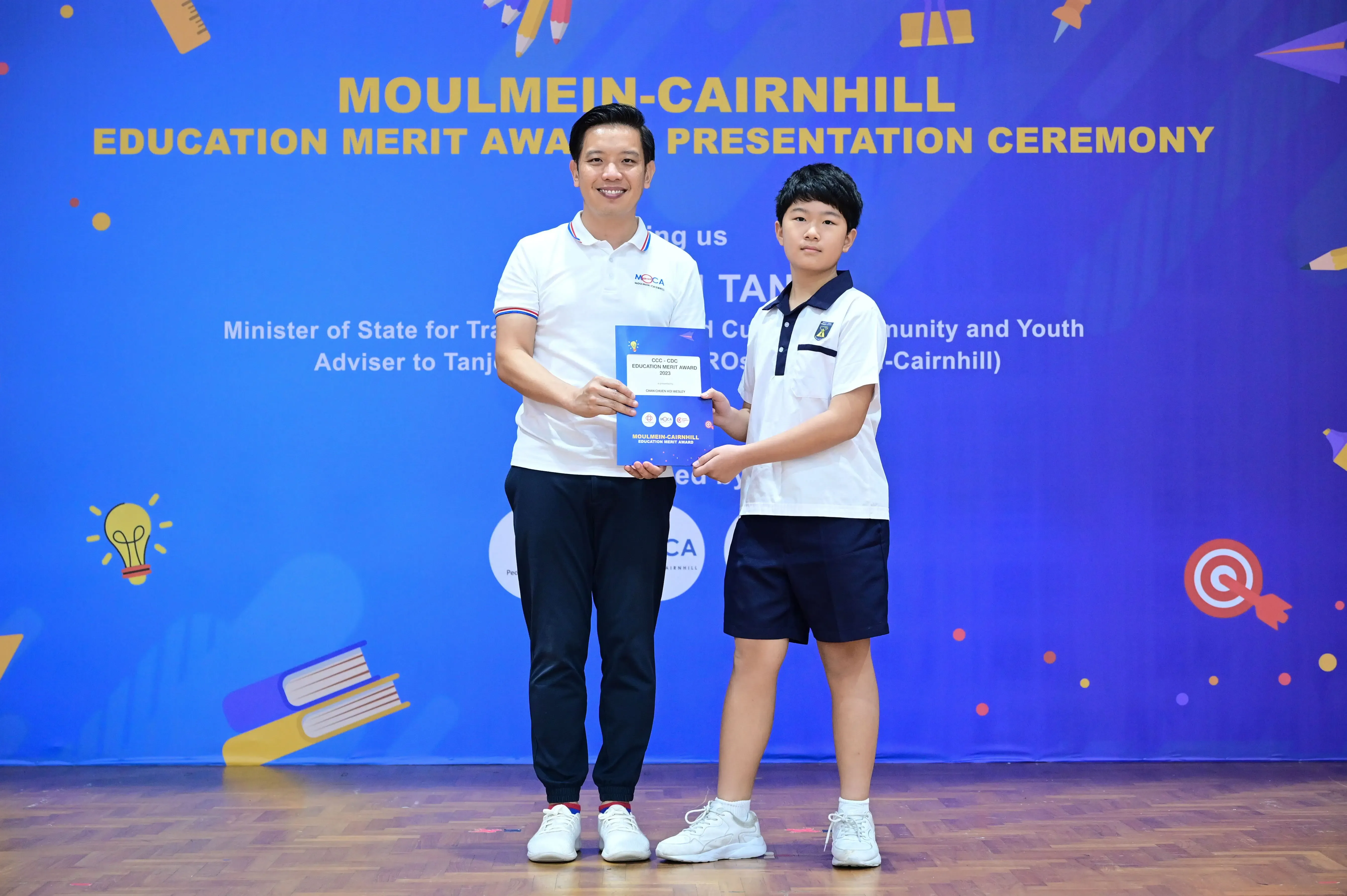 An adult and a young student hold a certificate together at the Moulmein-Cairnhill Education Merit Award Ceremony.