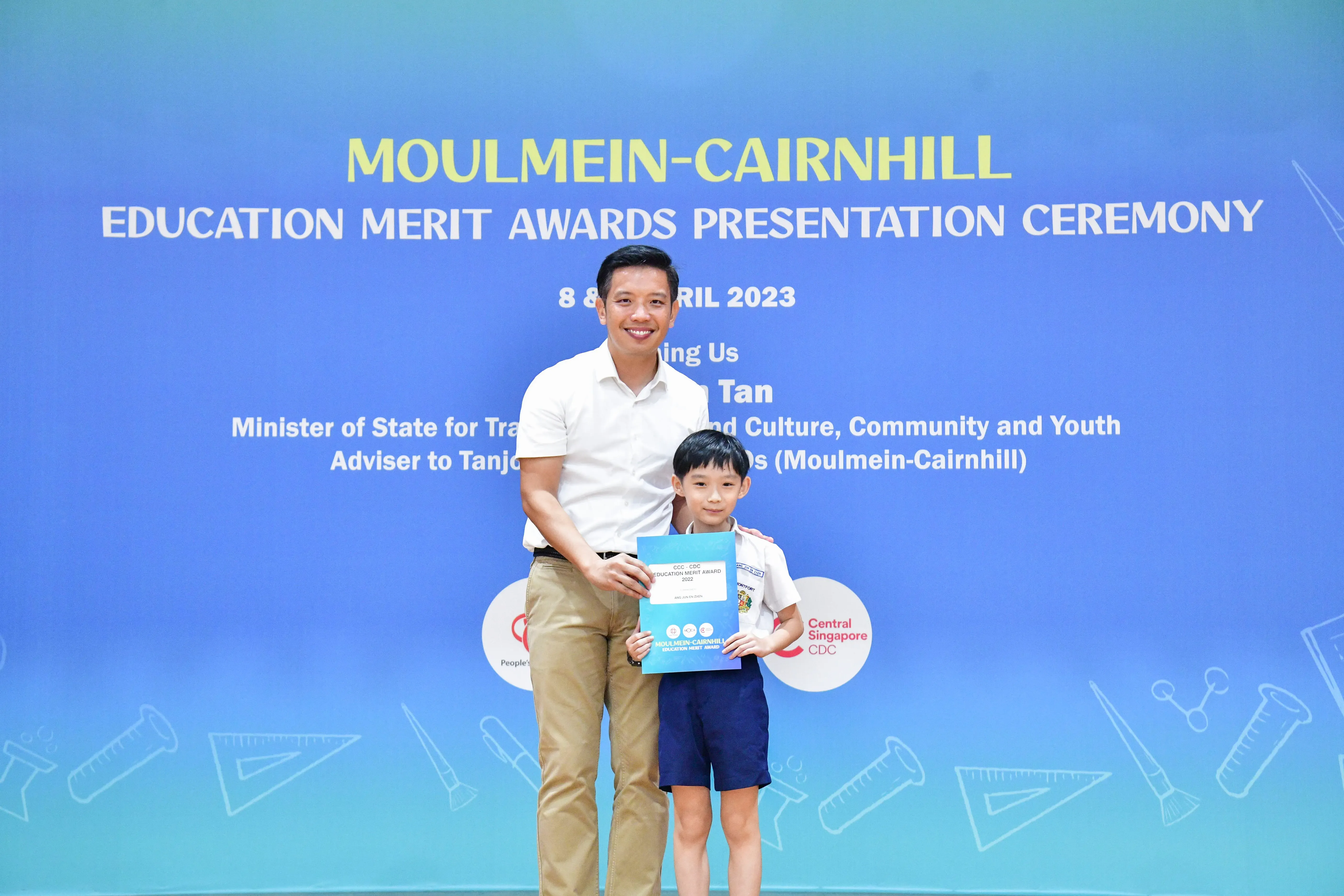 A man and young boy hold a certificate in front of a Moulmein-Cairnhill Education Merit Awards banner dated April 2023.