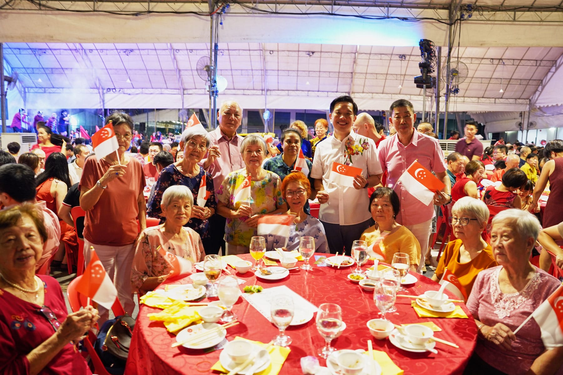A group of elderly and middle-aged people holding Singapore flags at a festive banquet under a large tent.