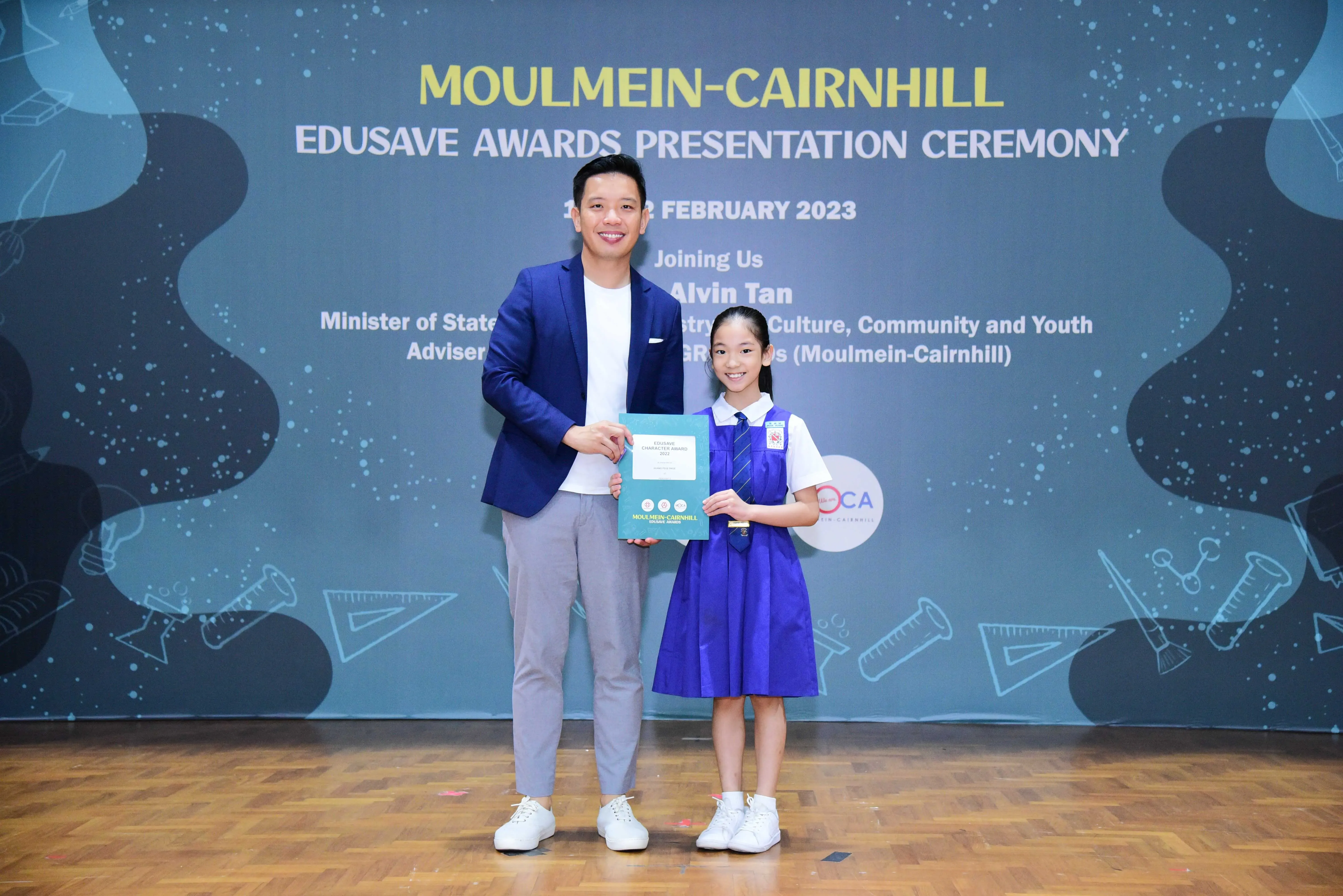 A man in a blue blazer and a young girl in a school uniform hold an award at the Moulmein-Cairnhill Edusave Awards Ceremony 2023.