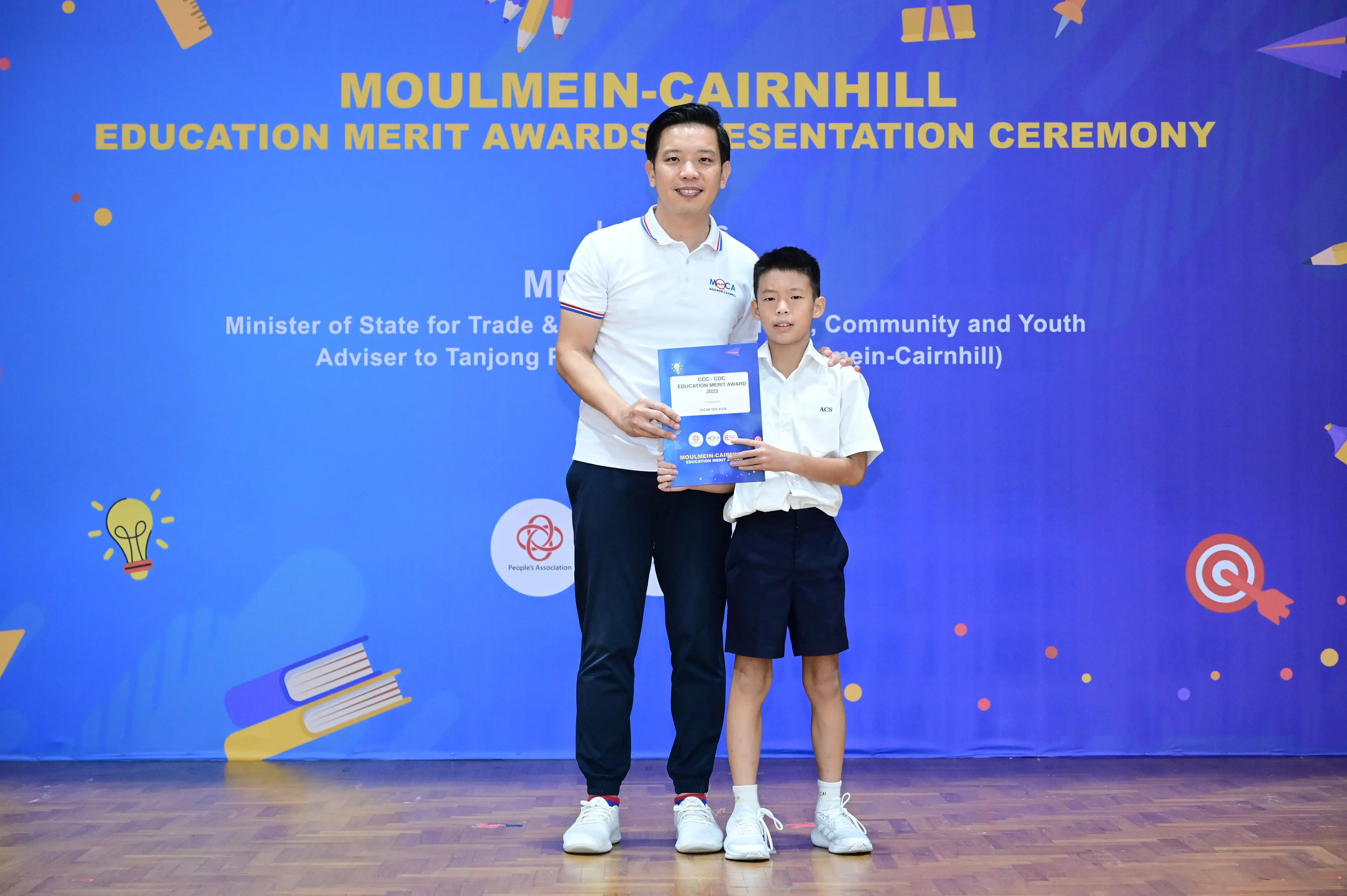 A man and a young boy hold a certificate together in front of a Moulmein-Cairnhill Education Merit Awards banner.