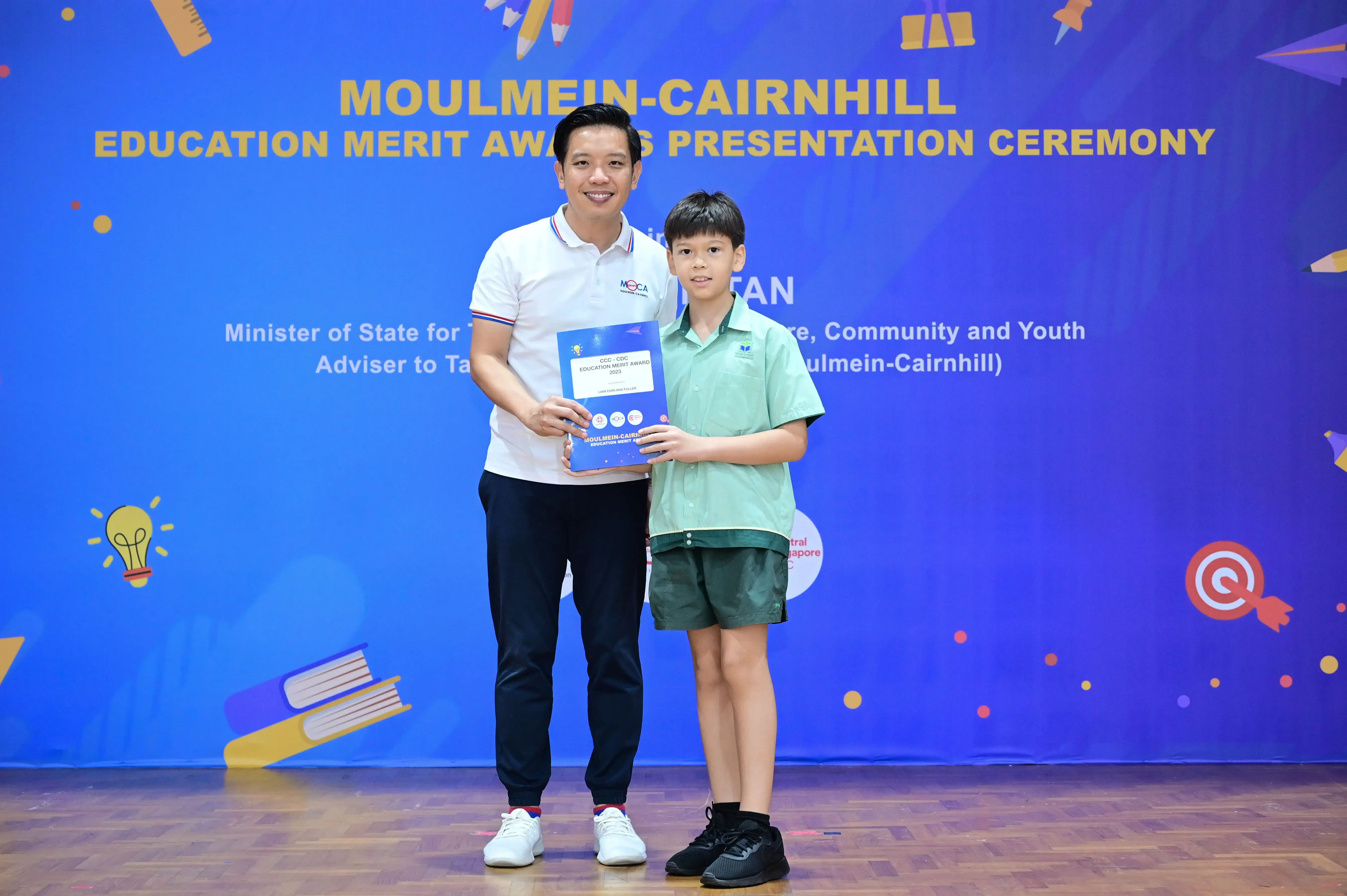 A man and a young boy hold a blue award folder at the Moulmein-Cairnhill Education Merit Awards Presentation Ceremony.