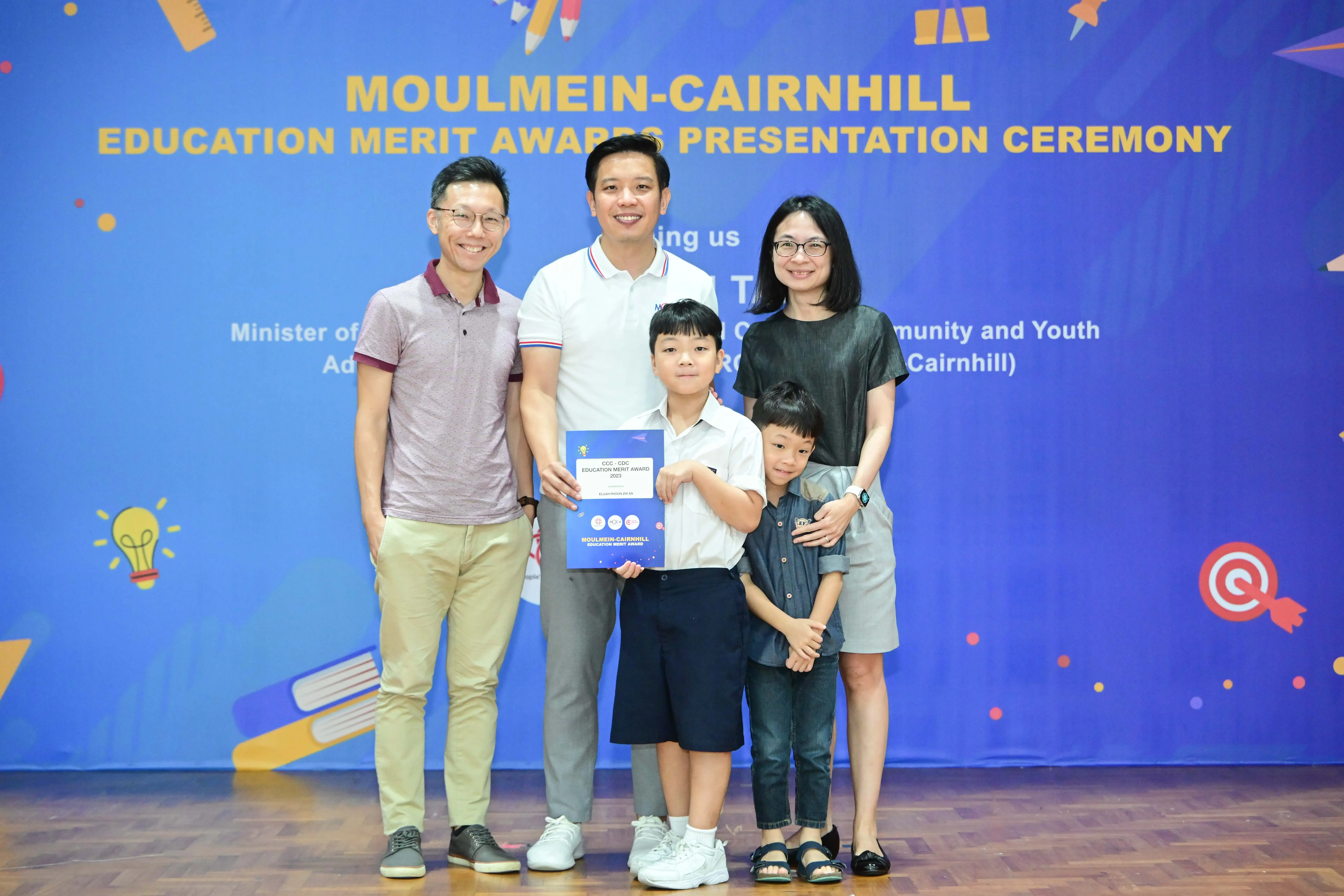 Five people pose at the Moulmein-Cairnhill Education Merit Awards ceremony, with a boy holding a blue certificate.