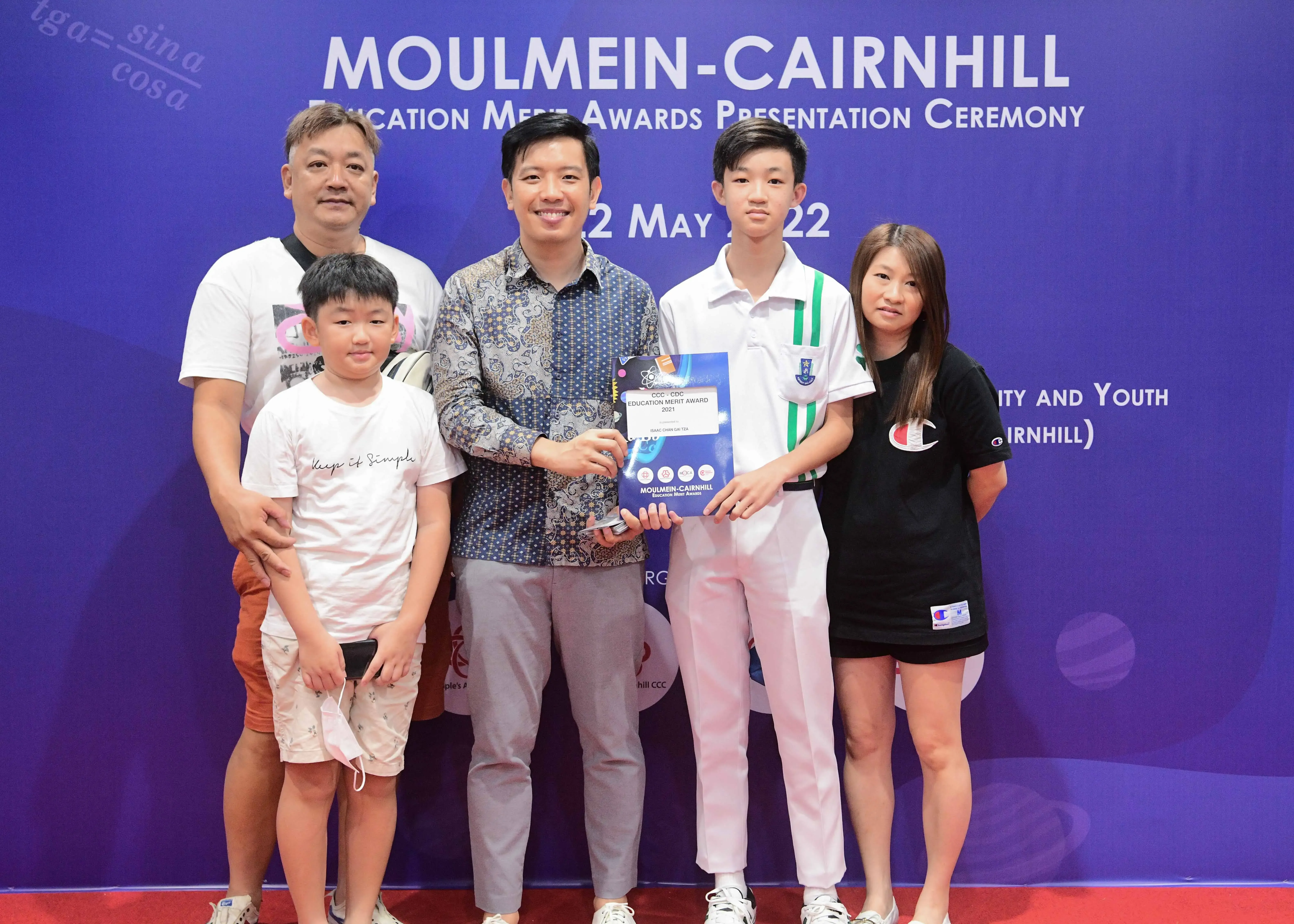 Five people pose in front of a Moulmein-Cairnhill Education Merit Awards Presentation Ceremony banner dated May 2022.