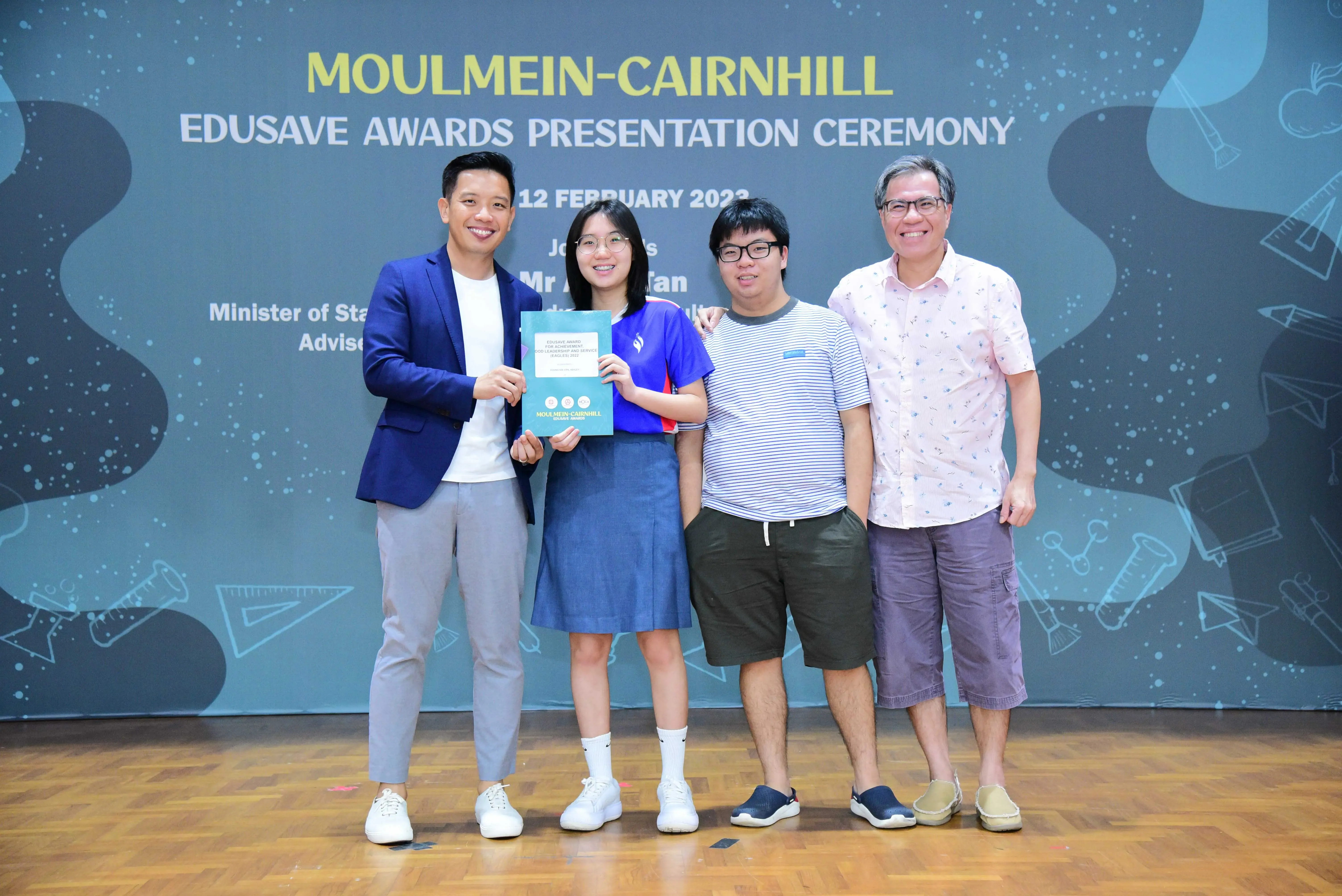 Four people smile at the Moulmein-Cairnhill Edusave Awards Presentation Ceremony, with a girl holding an award certificate.