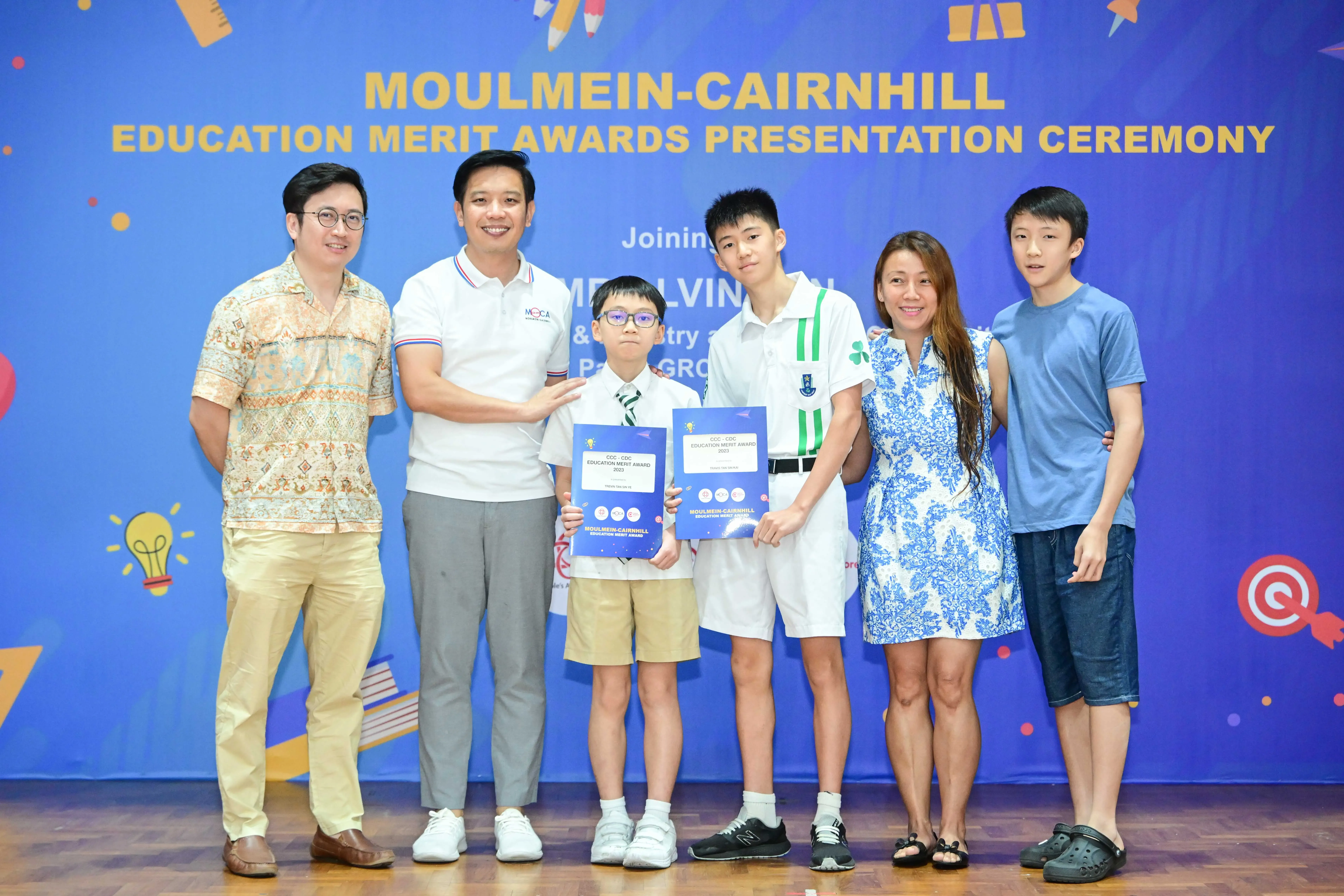 Six people pose at the Moulmein-Cairnhill Education Merit Awards ceremony, two boys holding blue award certificates.
