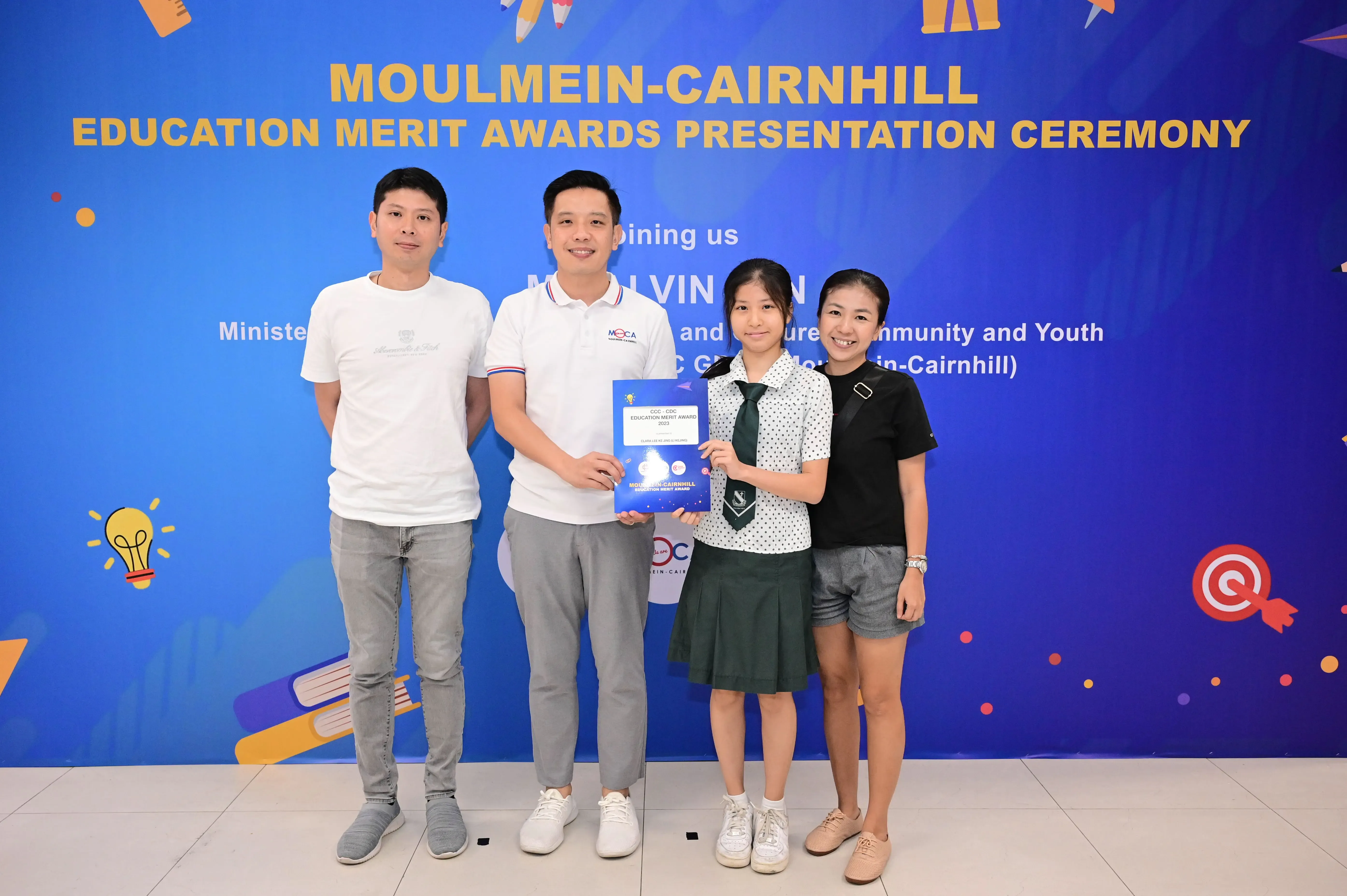 Four people pose before a Moulmein-Cairnhill Education Merit Awards banner, with a student holding a blue certificate.