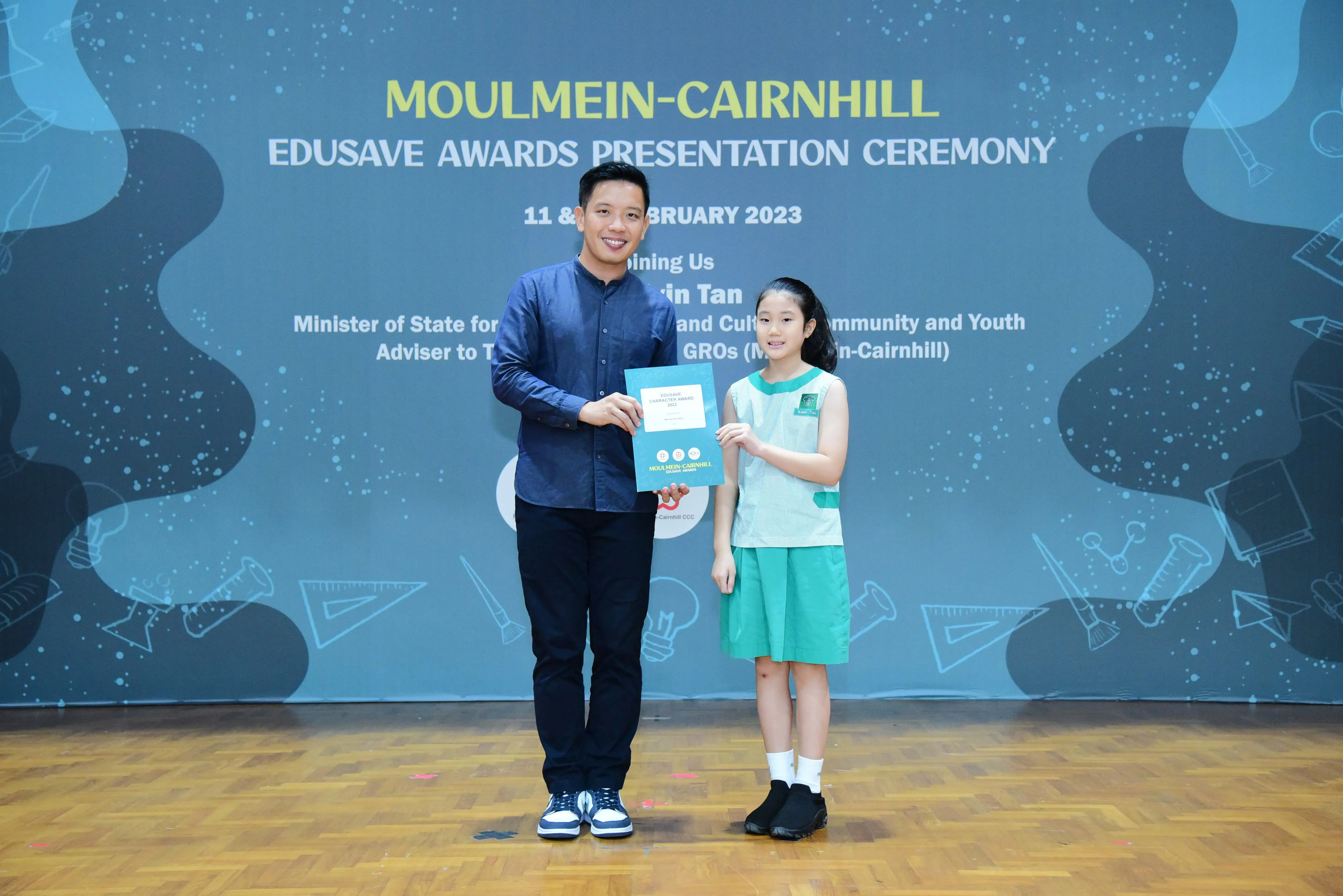 A man and a young girl hold a certificate together on stage at the Moulmein-Cairnhill Edusave Awards Ceremony 2023.