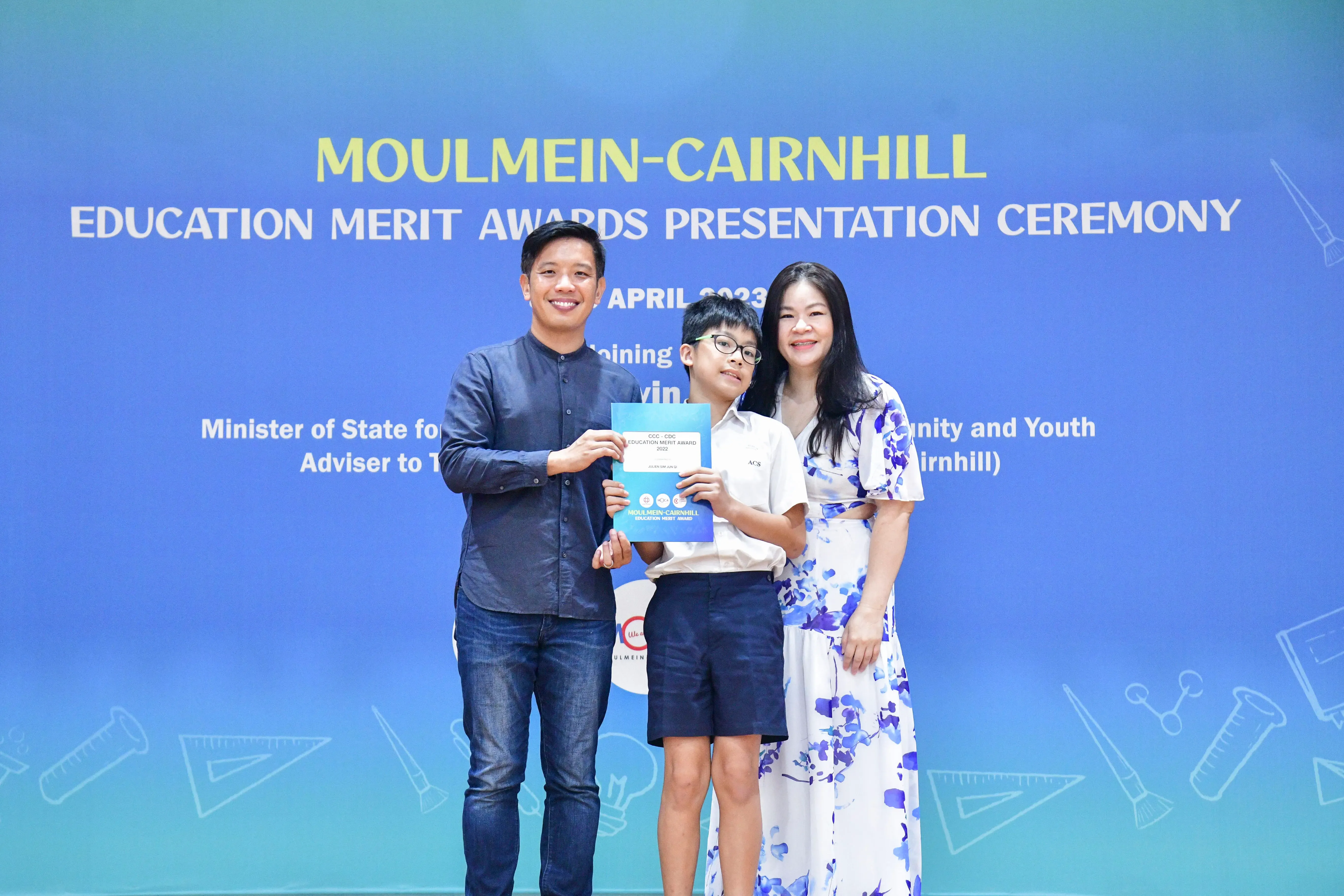 A boy holding an award certificate stands between two smiling adults at the Moulmein-Cairnhill Education Merit Awards 2023.