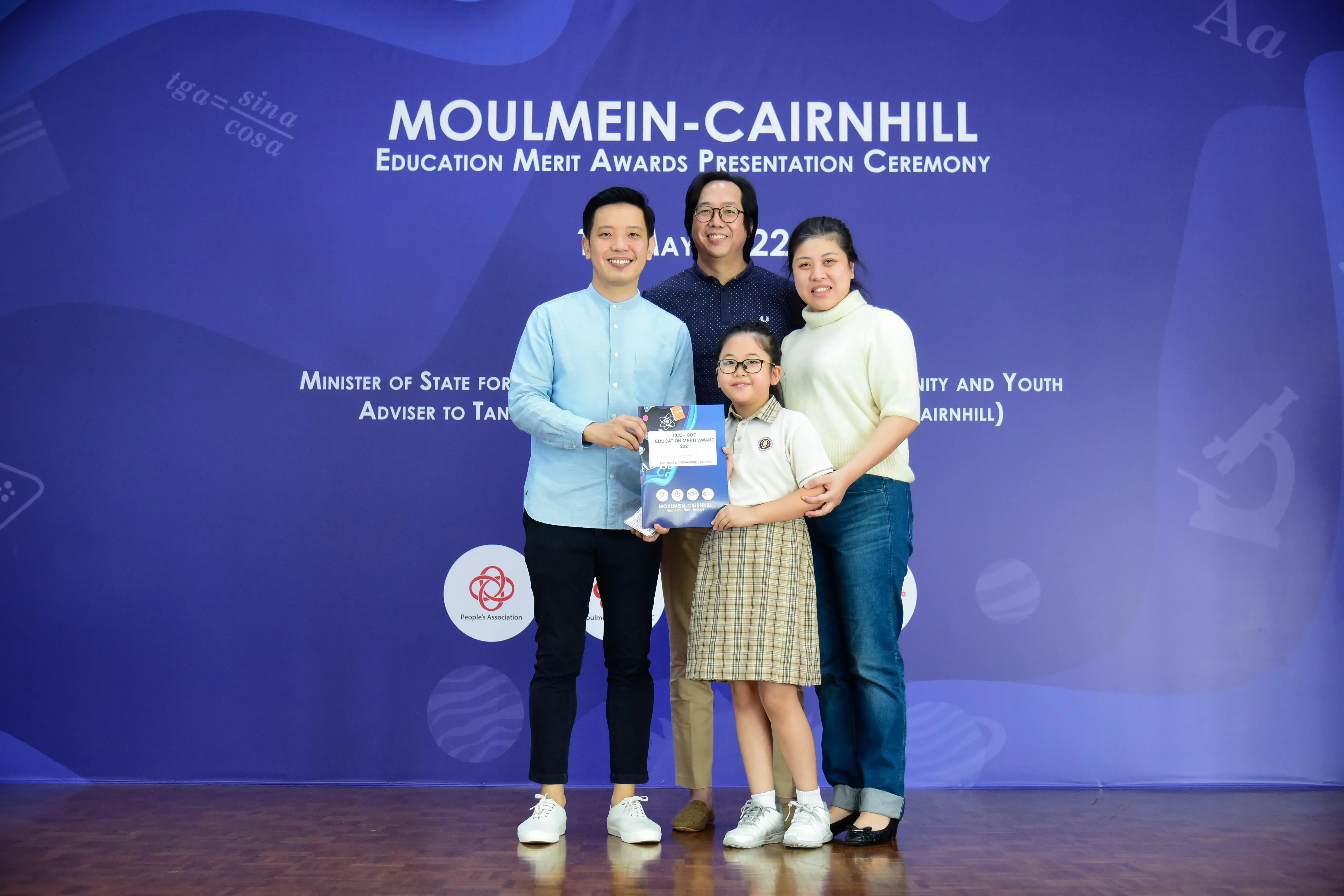 Four people pose at the Moulmein-Cairnhill Education Merit Awards ceremony, with a young girl holding an award certificate.