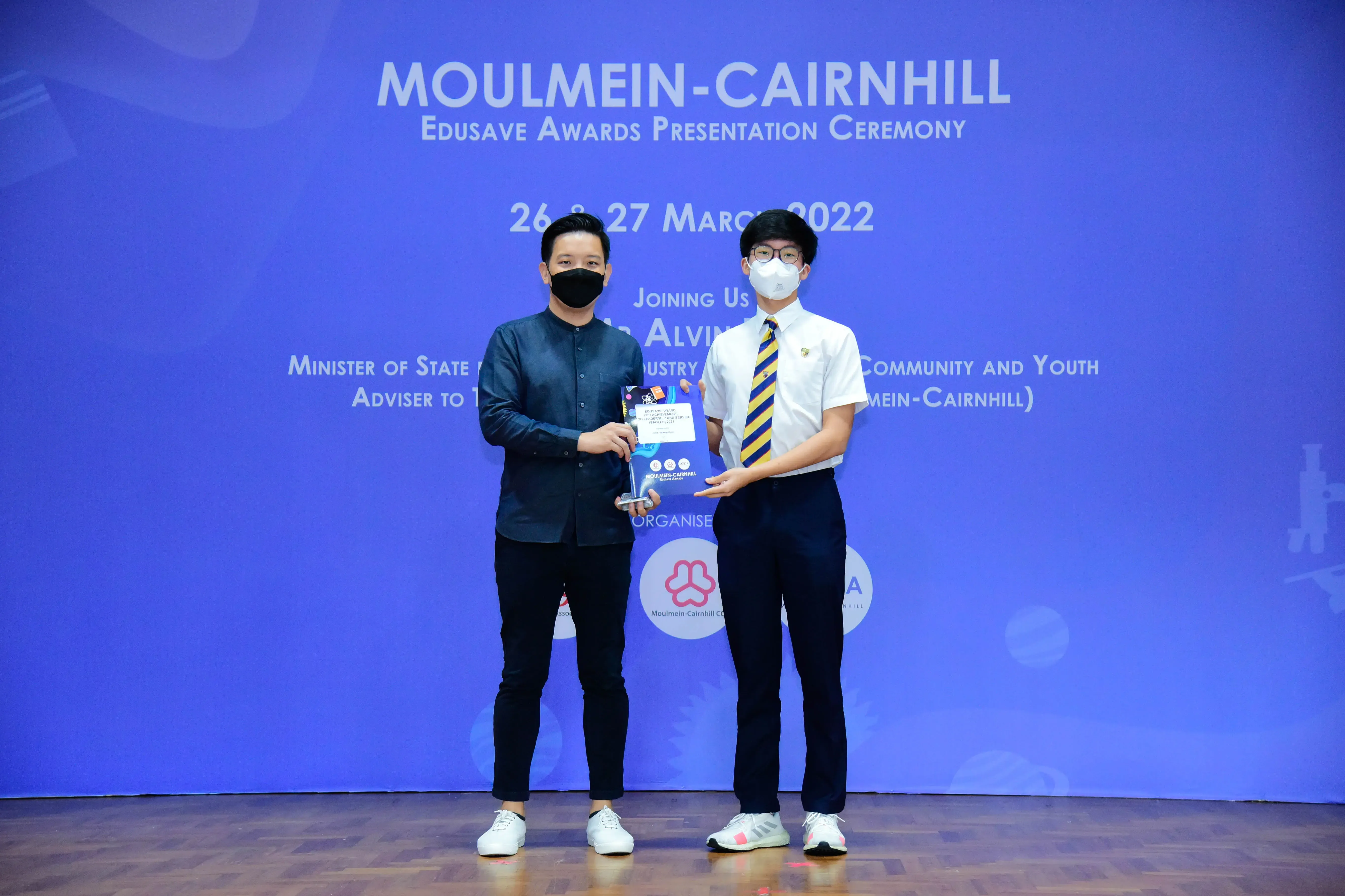 Two masked men hold an award at the Moulmein-Cairnhill Edusave Awards Presentation Ceremony, March 2022.