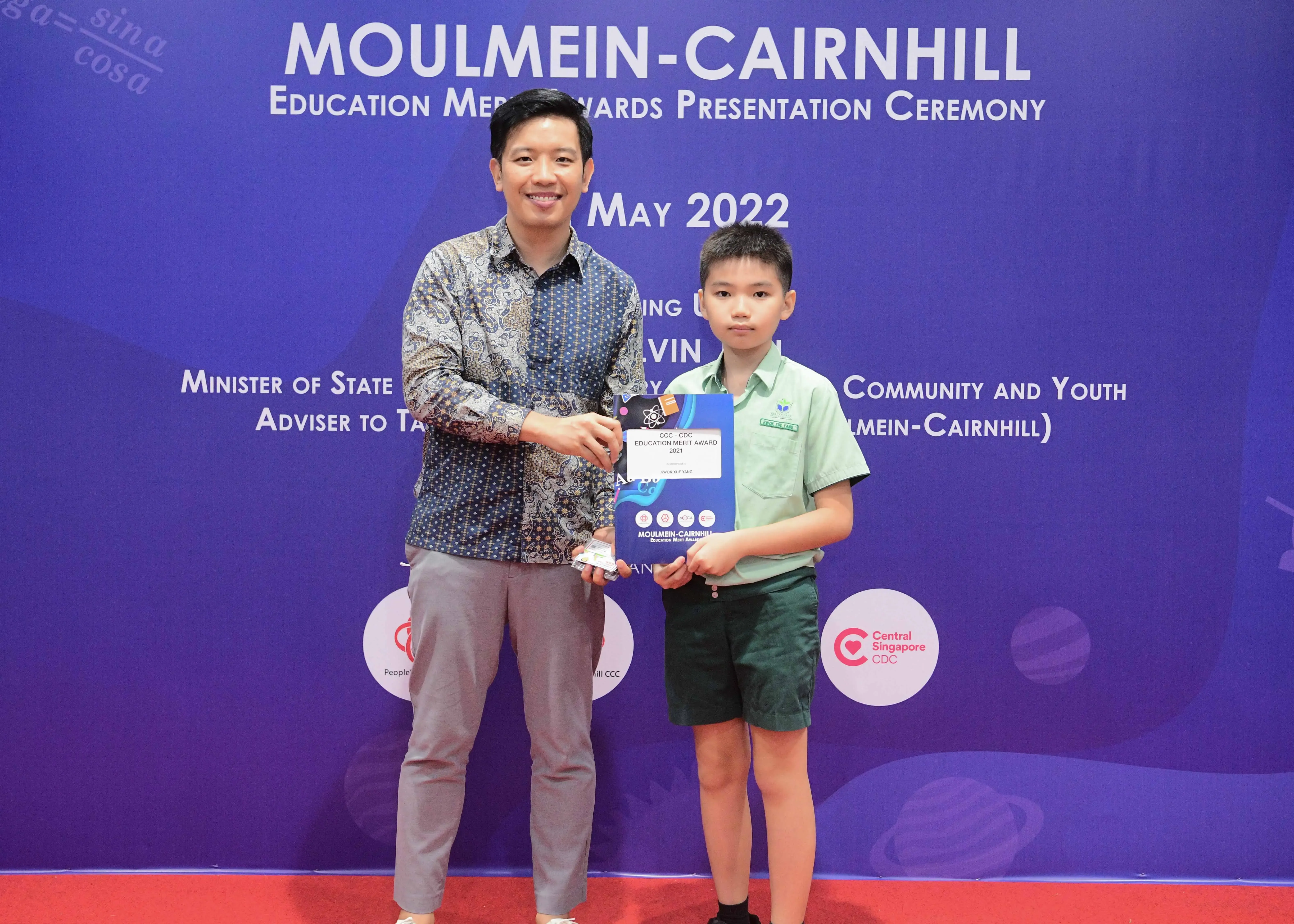 An adult and a young boy hold an award together at the Moulmein-Cairnhill Education Merit Awards Ceremony, May 2022.