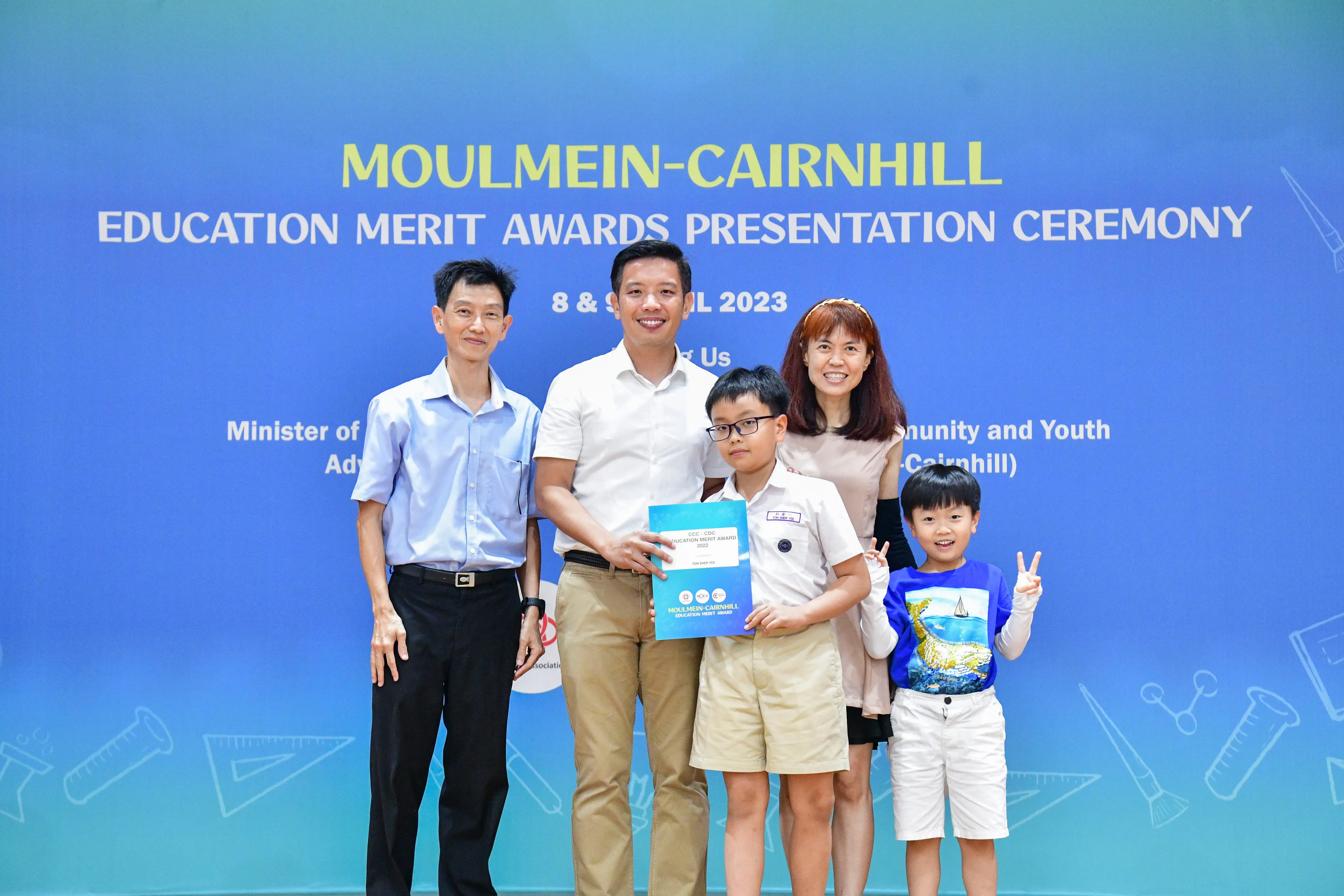 Five people pose in front of a blue banner for the Moulmein-Cairnhill Education Merit Awards Ceremony 2023.